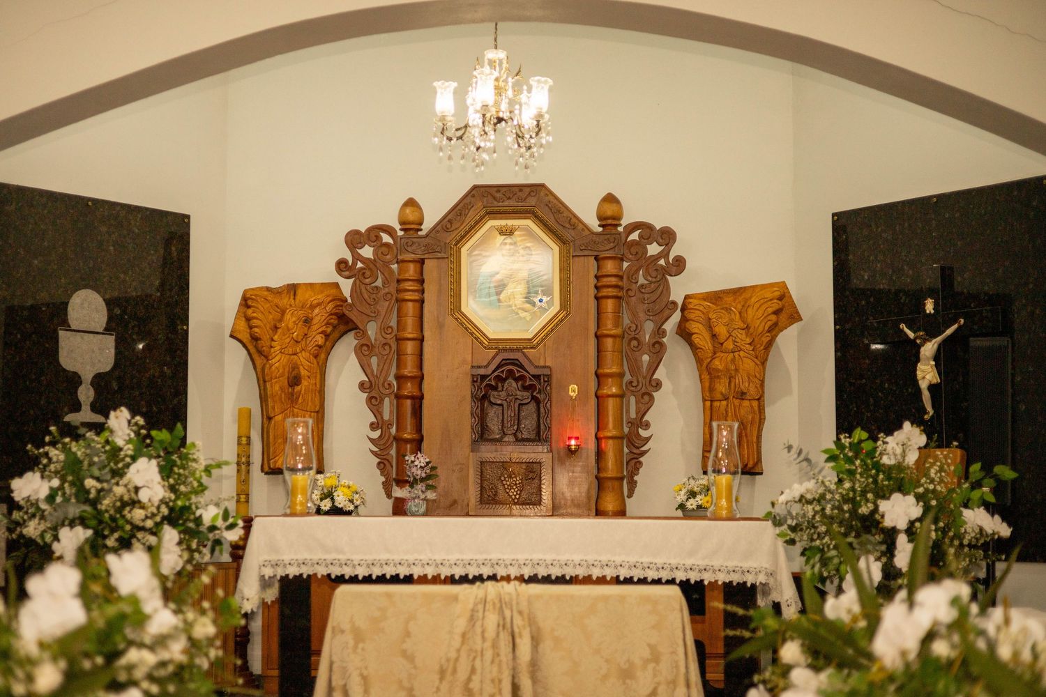 Ornate wooden church altar with white floral arrangements, chandelier, and crucifix in a Catholic chapel.
