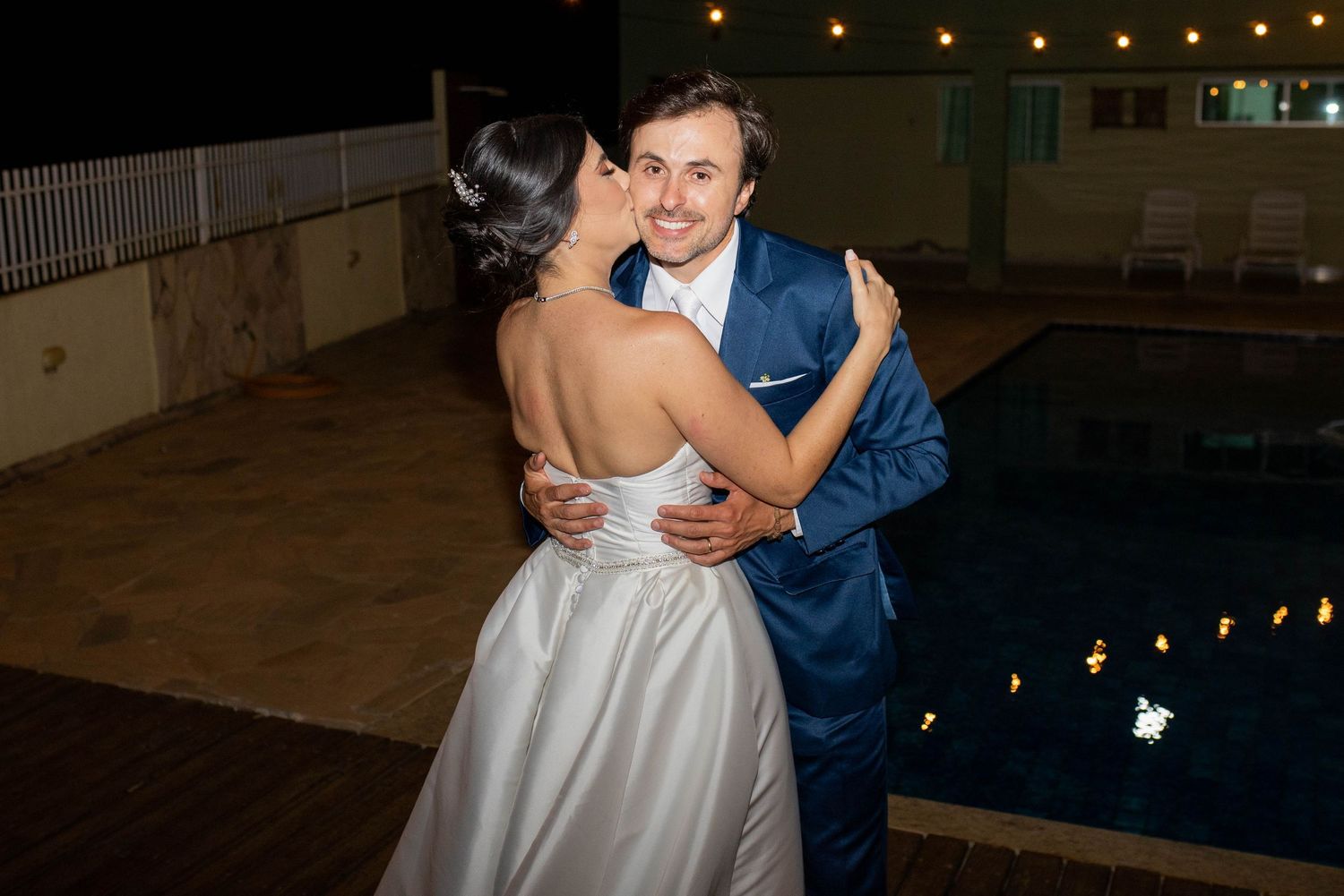 Bride and groom share a romantic dance outdoors at night beside a pool with string lights at their wedding reception.