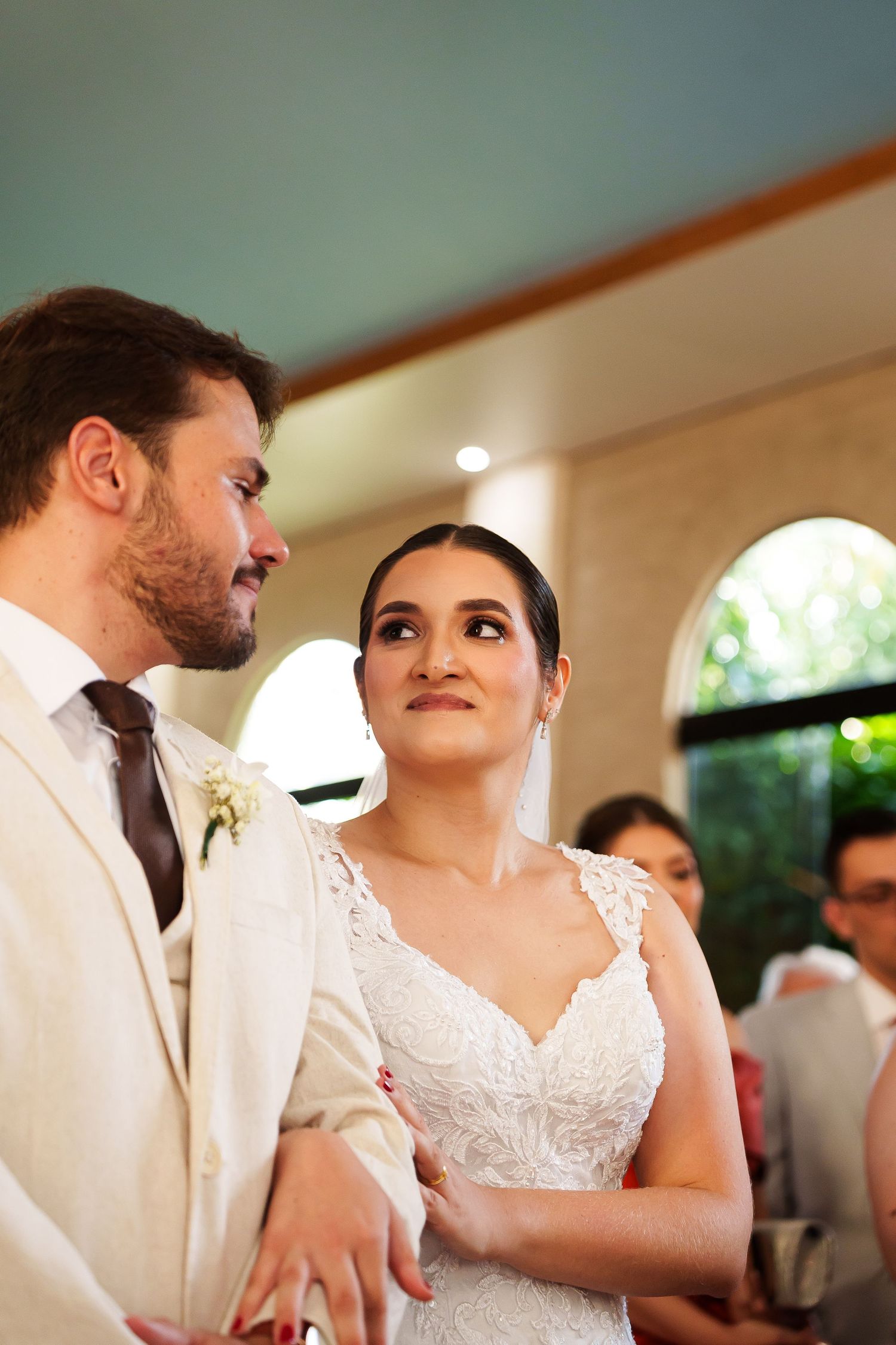 A bride in a white lace gown gazes lovingly at her groom in a beige suit during their wedding ceremony.