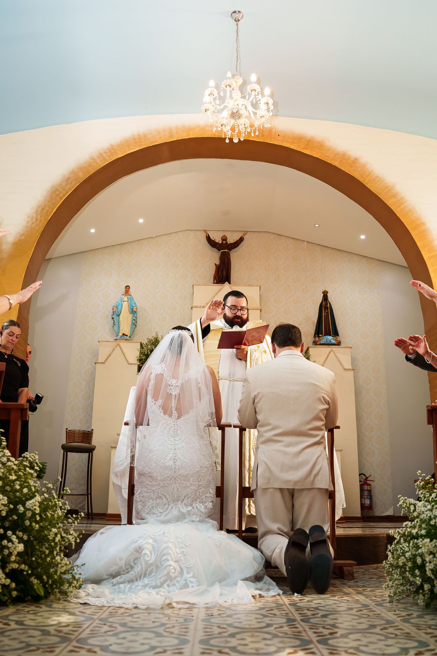 Bride and groom kneeling at church altar during Catholic wedding ceremony with priest blessing the couple.