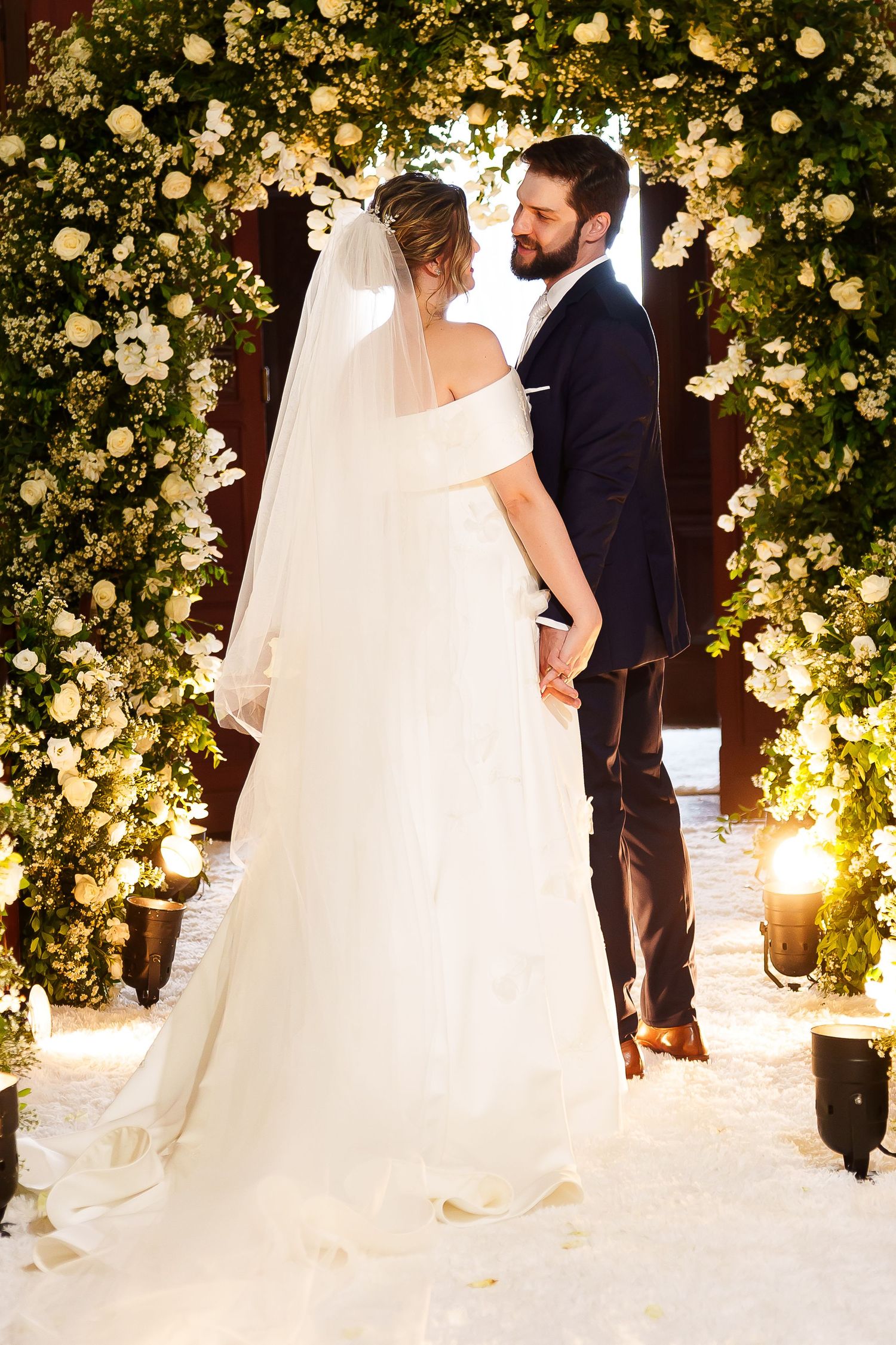 Bride and groom holding hands at floral wedding altar adorned with white roses and greenery in romantic candlelit ceremony.
