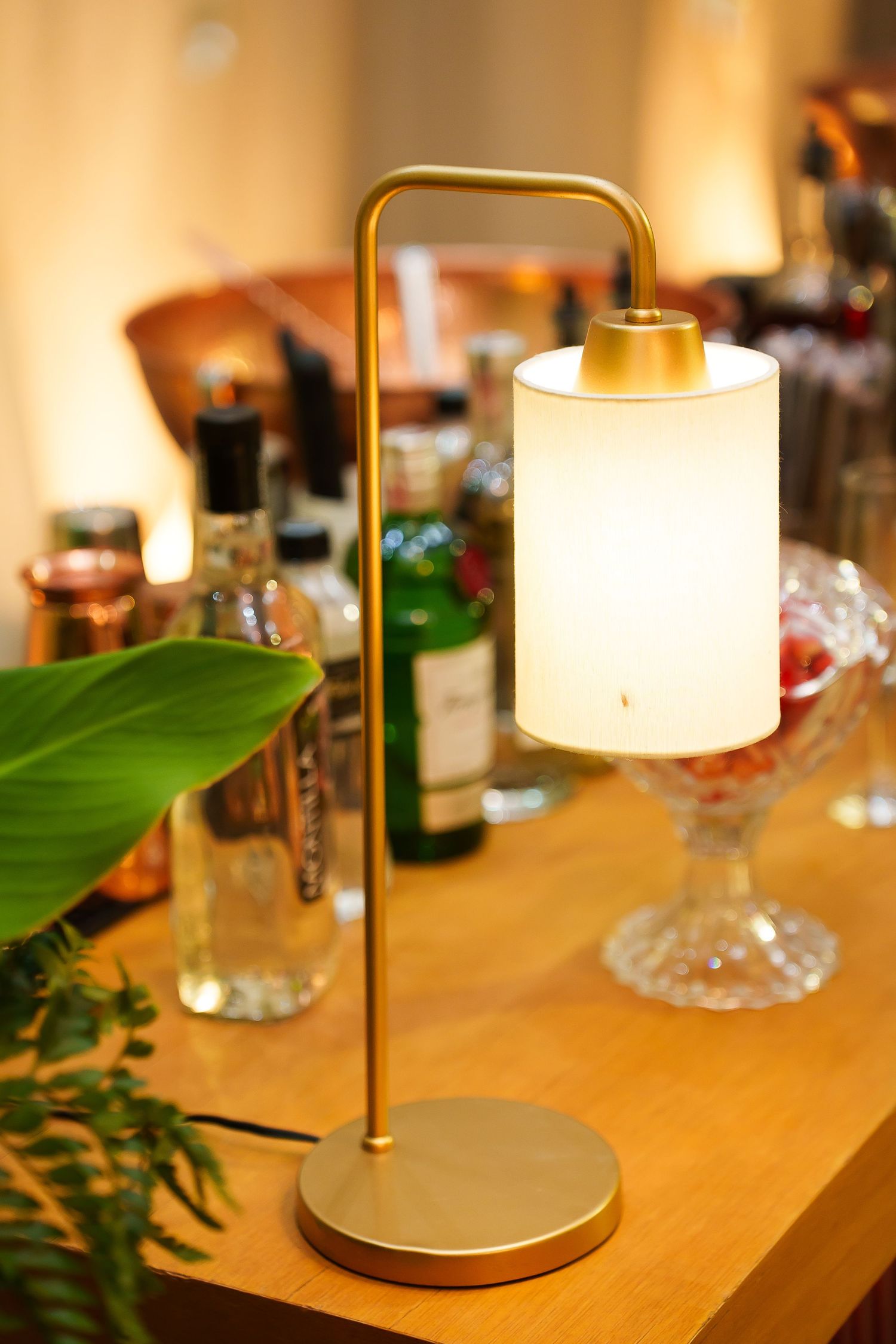 Gold brass table lamp with white cylindrical shade on wooden bar counter surrounded by bottles and glassware.