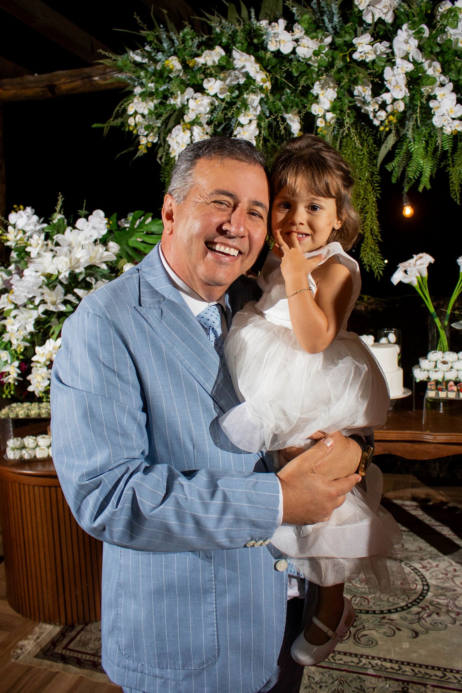 Smiling man in blue suit holding a little girl in white dress at an elegant floral event.