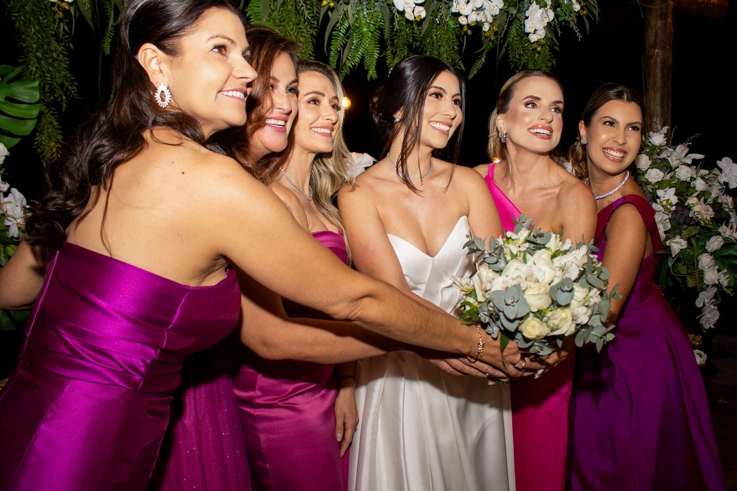 Bride in white gown holds bouquet surrounded by smiling bridesmaids in purple and pink dresses at wedding reception.
