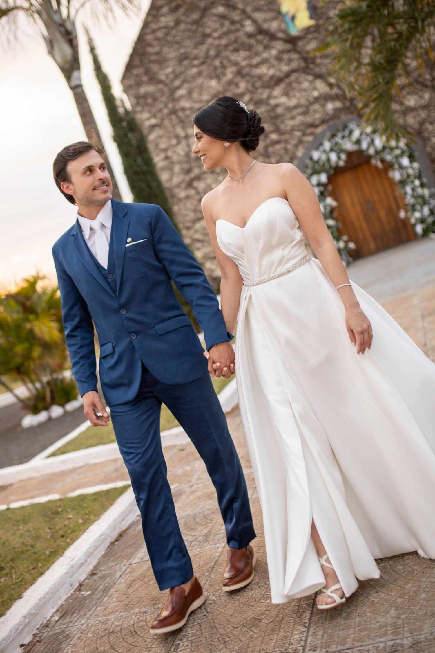 A bride in a white strapless gown and groom in a blue suit hold hands while walking outdoors at their wedding venue.