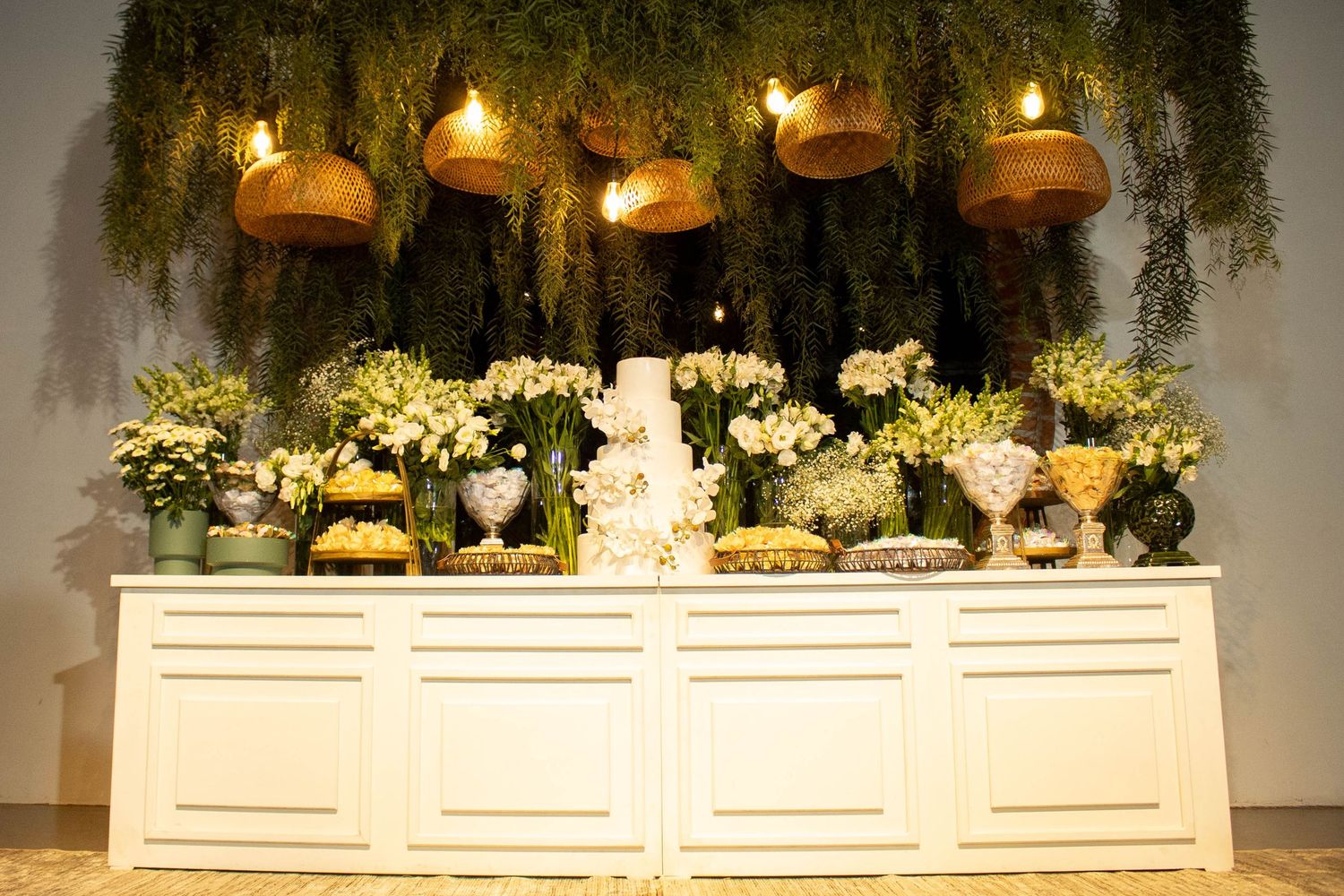 Elegant wedding dessert table with white cake, floral arrangements, and hanging greenery with golden basket decorations.
