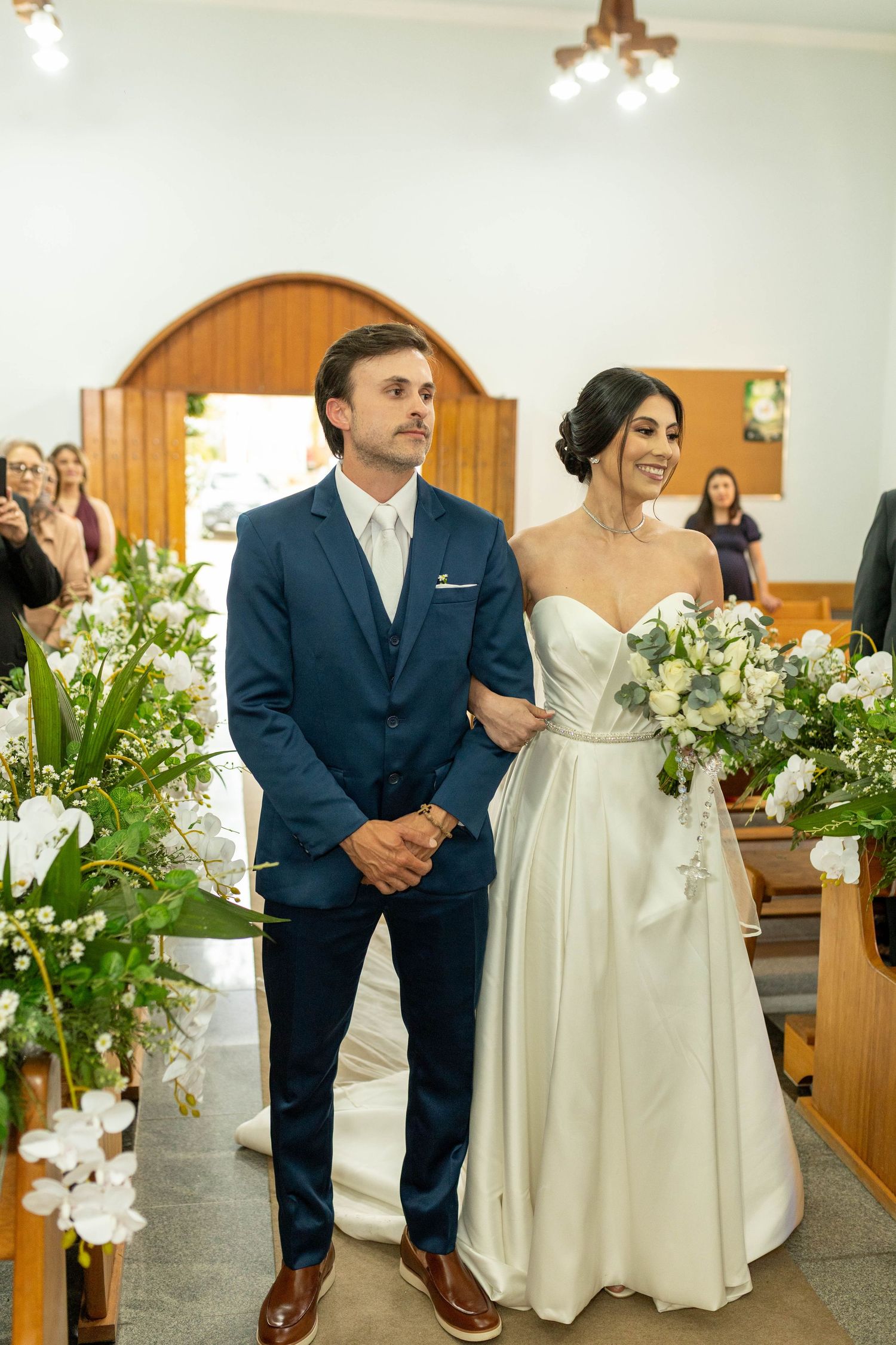 Bride in white gown holding bouquet stands with groom in navy suit at church wedding ceremony altar.