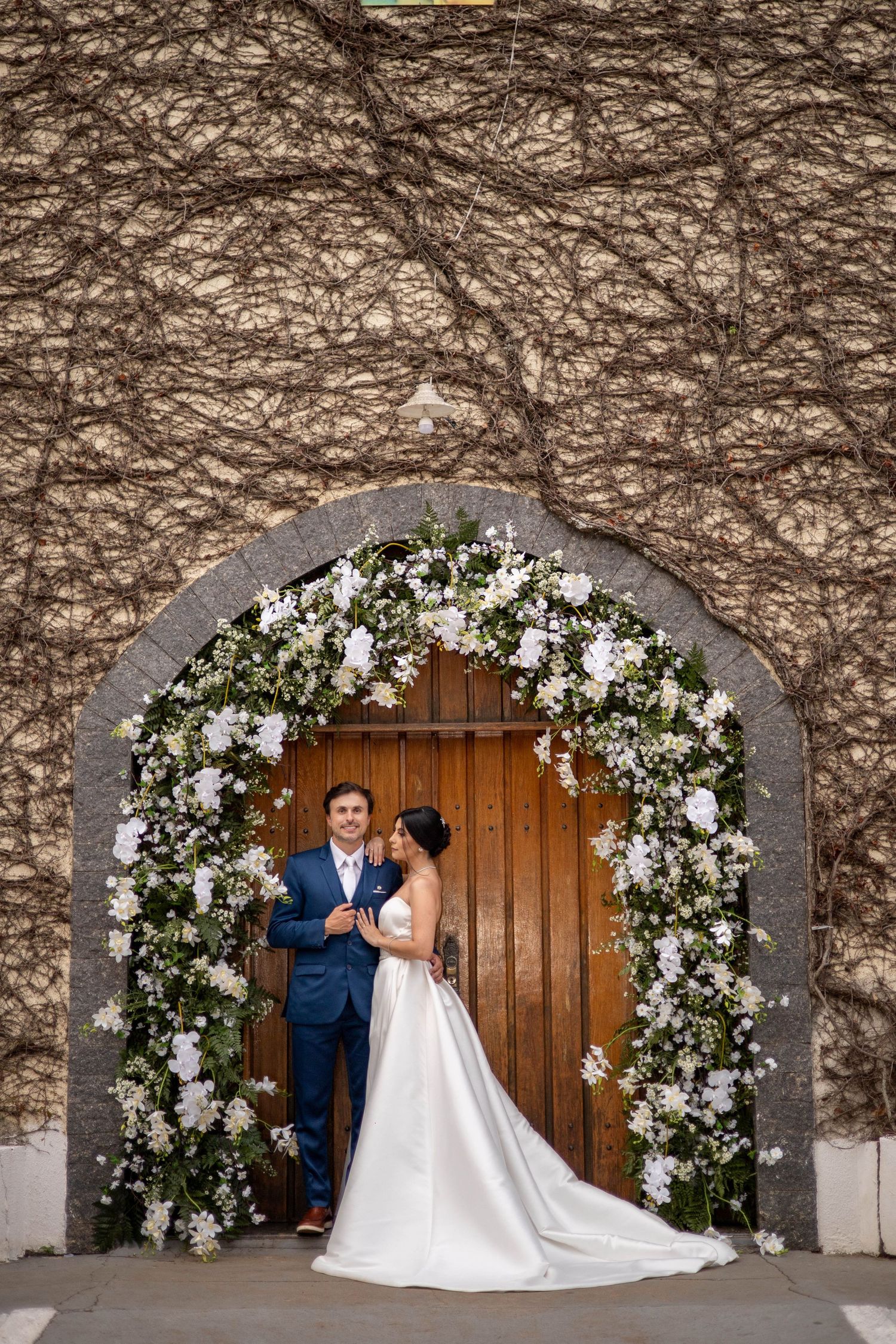 Bride and groom standing under a floral arch with white flowers at a wooden door covered in vines.