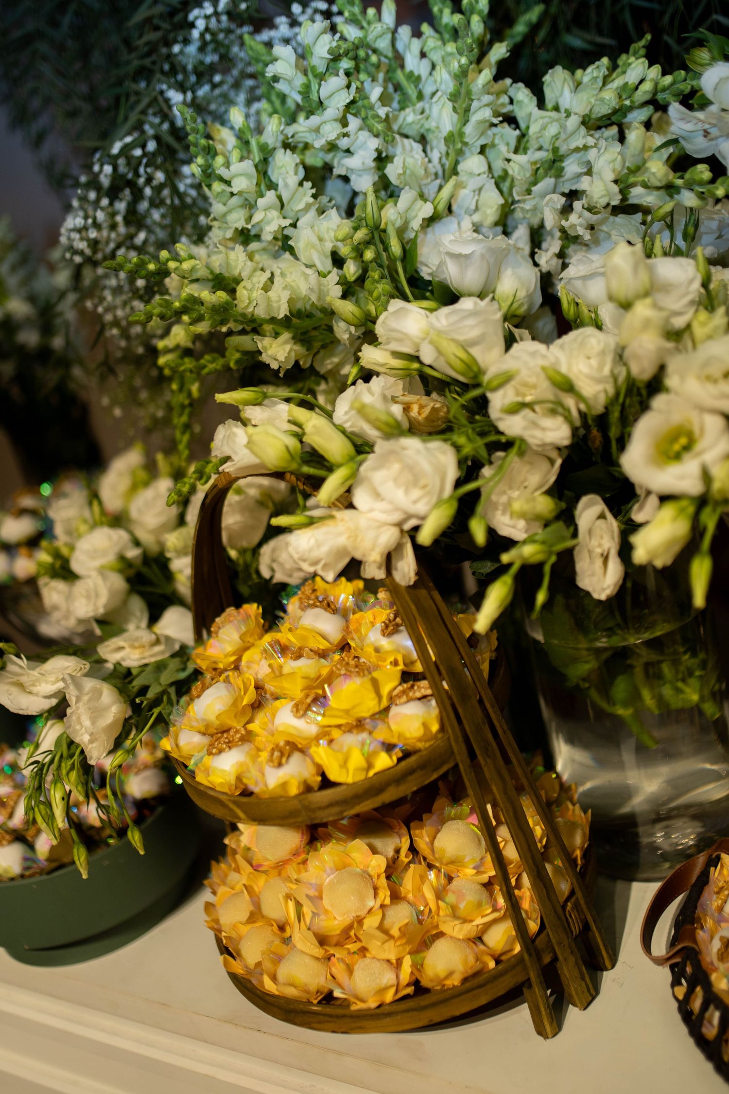 Two-tiered gold stand with yellow petit fours surrounded by white eustoma and delphinium floral arrangements on a white table.