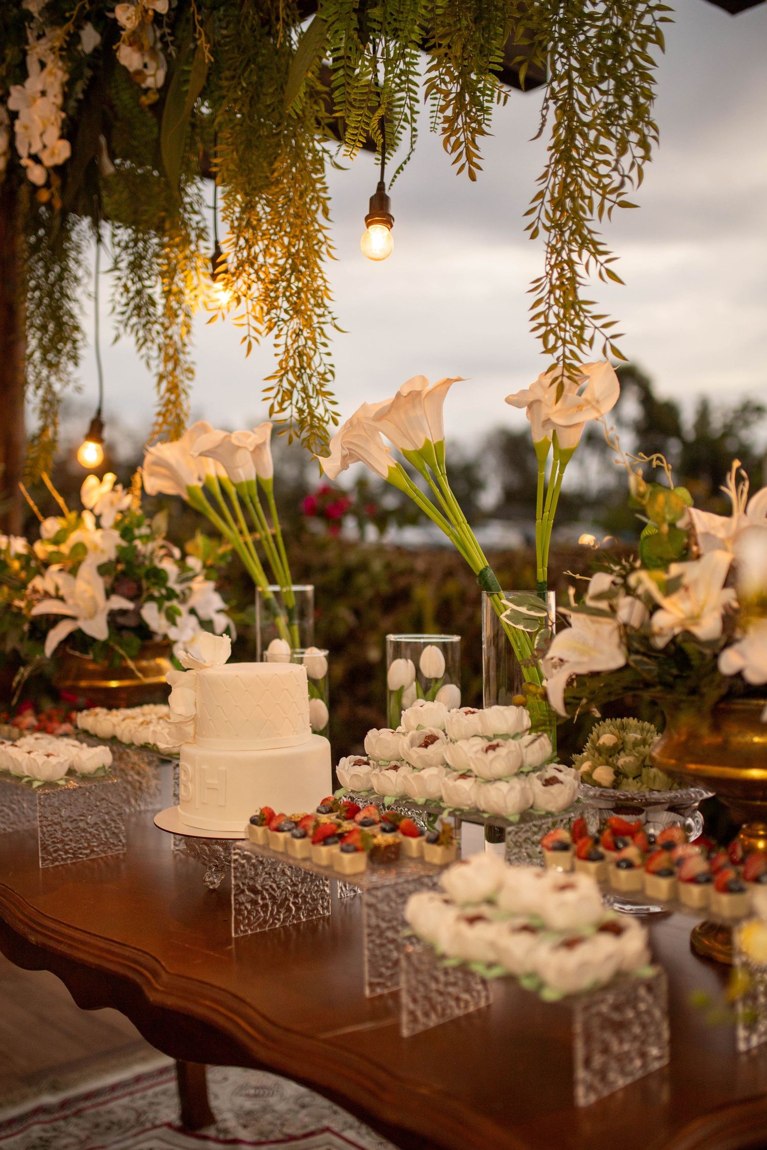 Elegant wedding dessert table with white tiered cake, floral arrangements, and assorted sweets under hanging greenery lights.