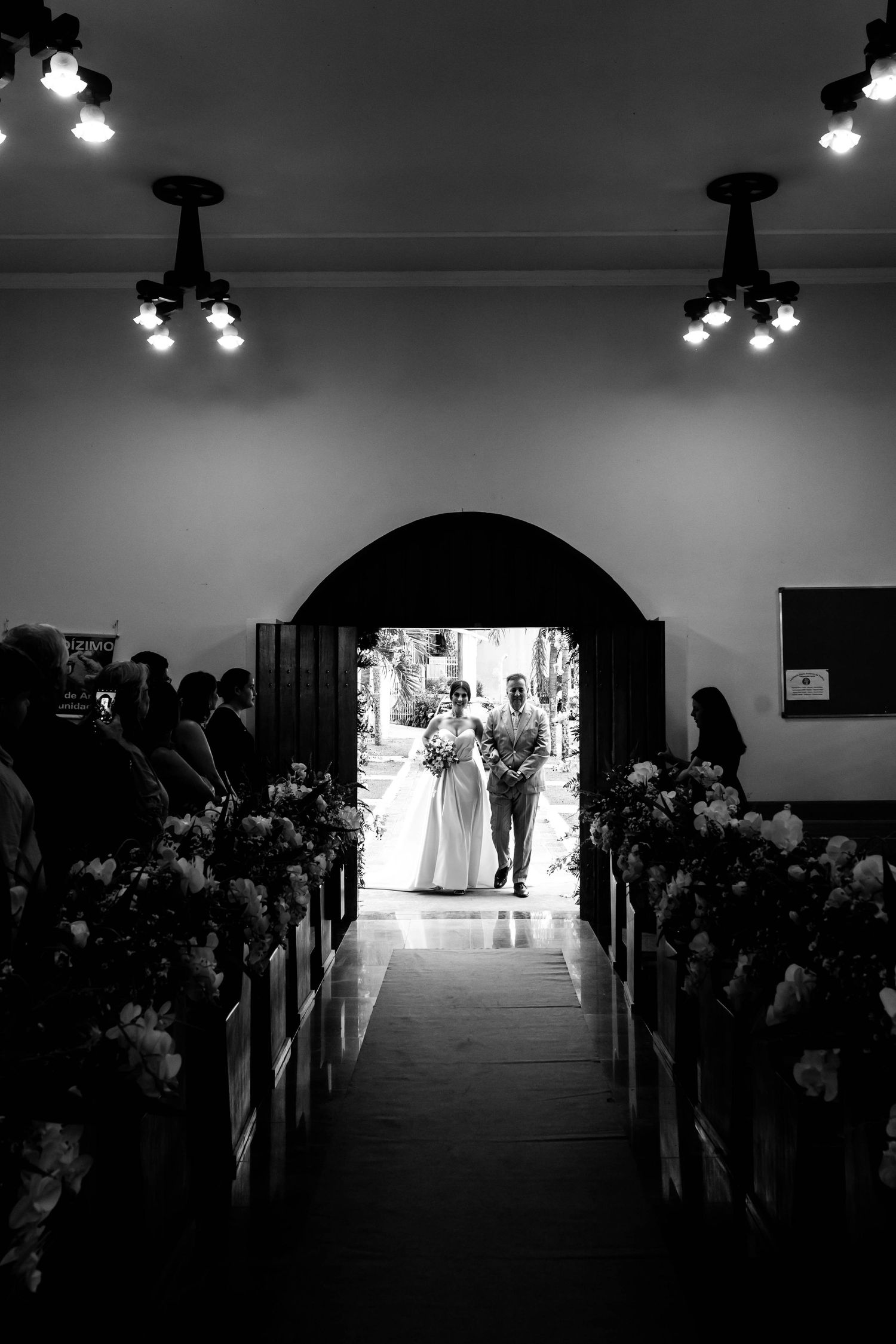 Black and white photo of a bride and groom entering a church aisle lined with flowers and guests during a wedding ceremony.