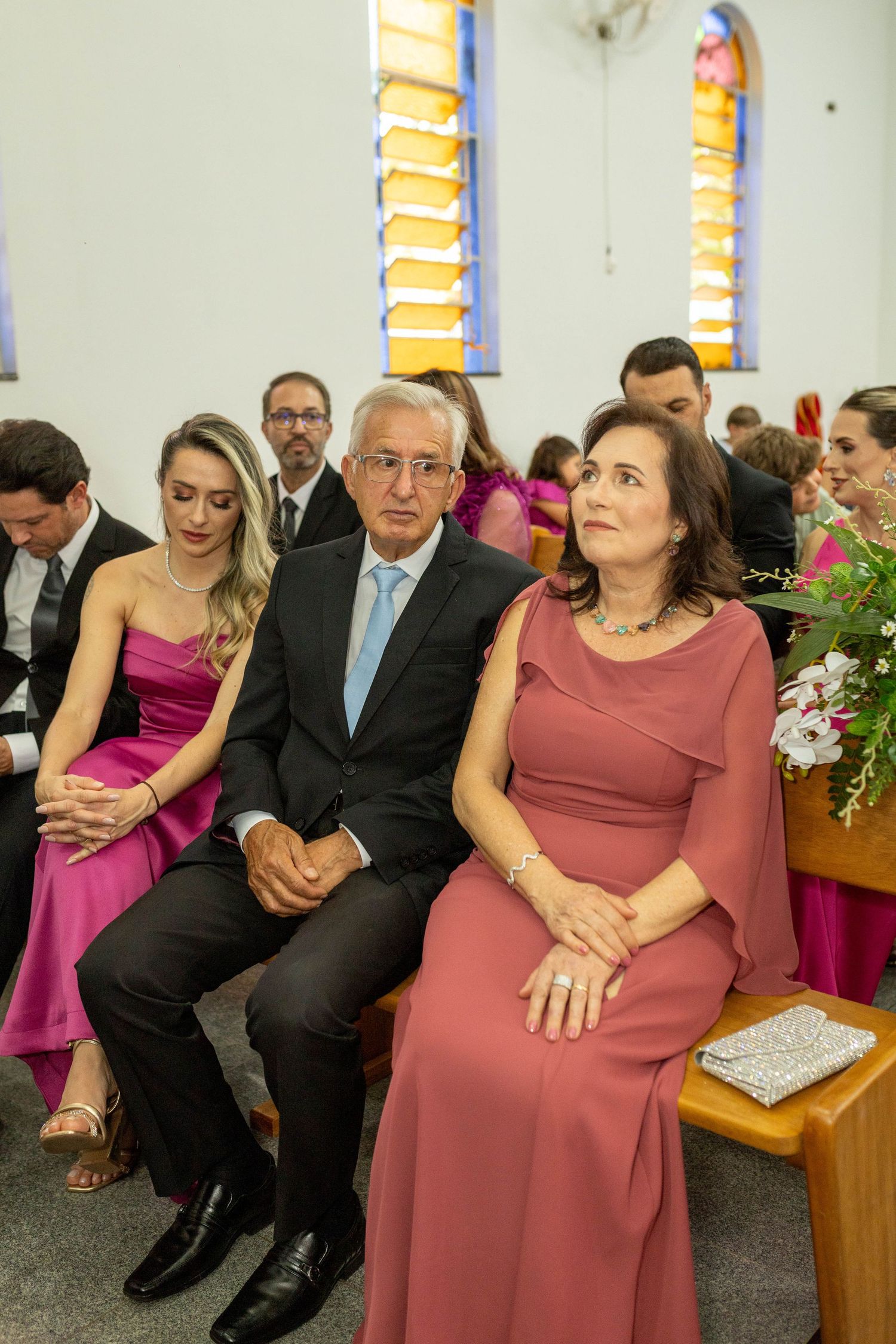 Elegantly dressed wedding guests seated in church pews, wearing pink and rose formal attire during a ceremony.