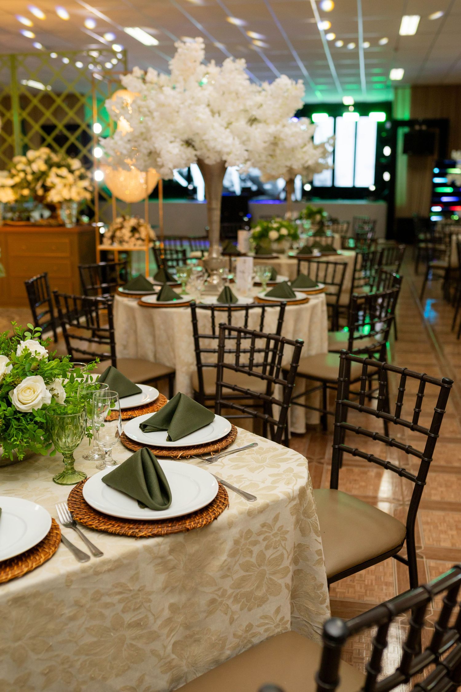 Elegant wedding reception tables with green napkins, wicker chargers, and tall white floral centerpieces in a decorated venue.
