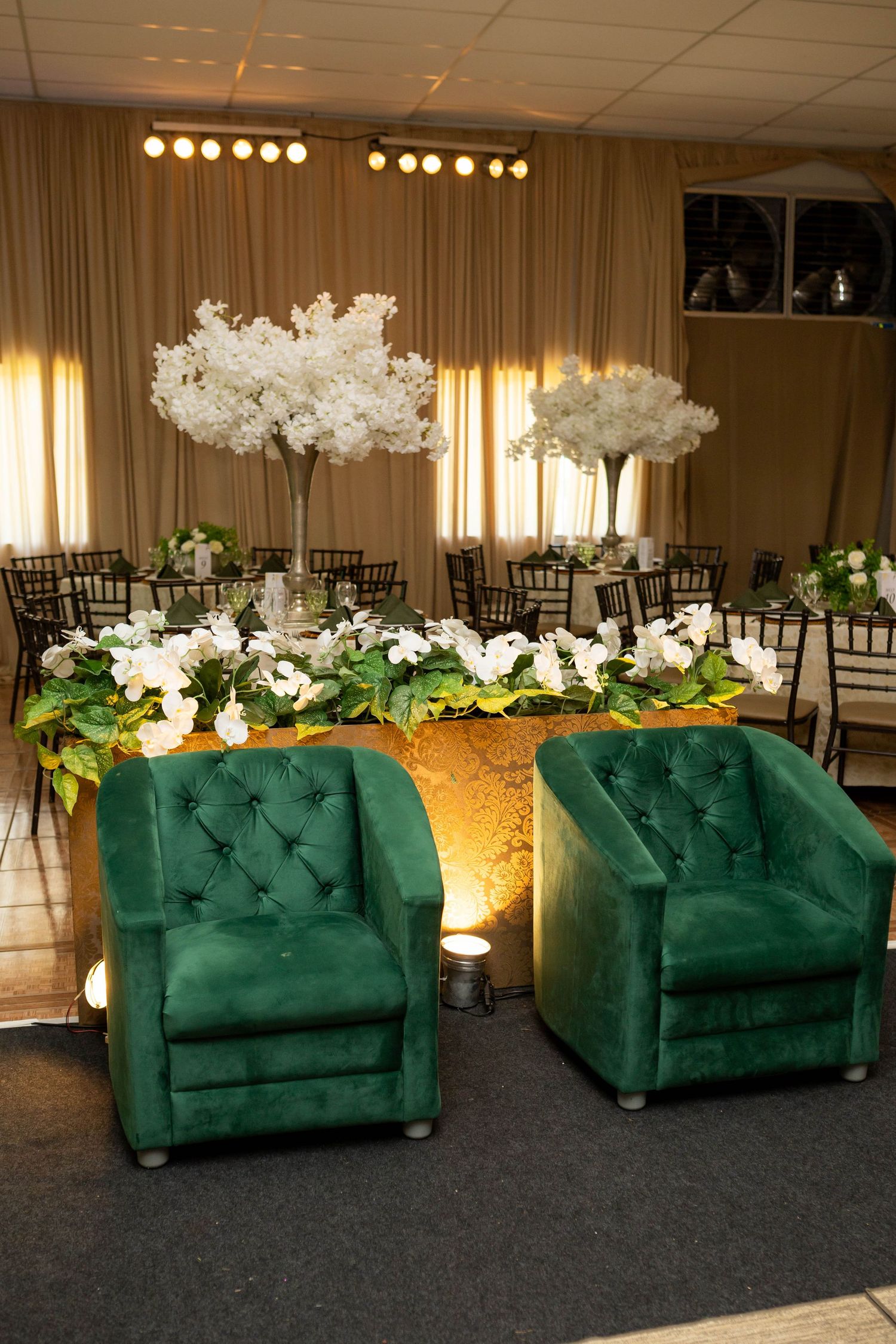 Two tufted emerald green velvet armchairs in front of a floral wedding sweetheart table with white cherry blossom centerpieces.