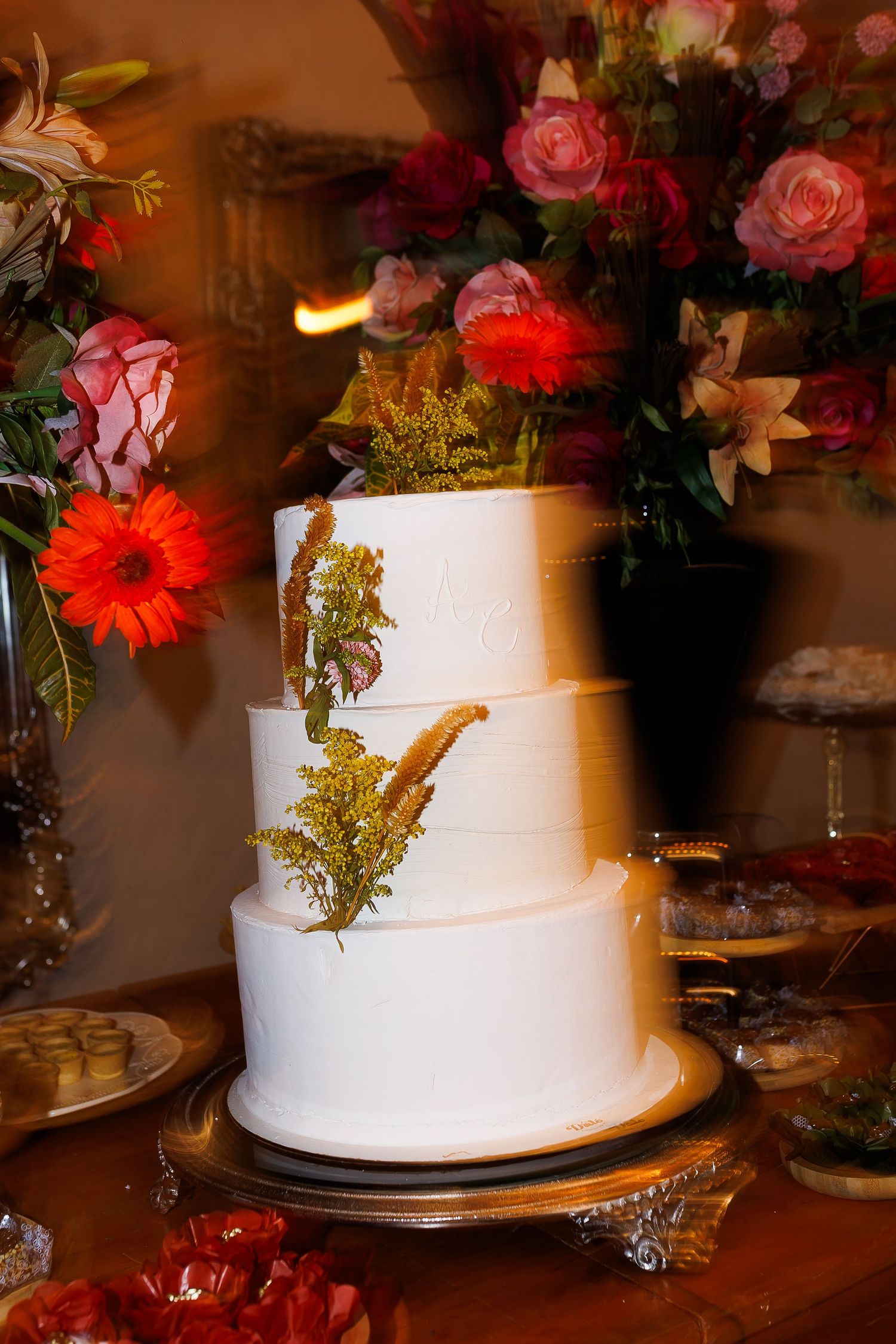 Elegant three-tier white wedding cake adorned with dried botanicals on a gold stand, surrounded by vibrant floral arrangements.