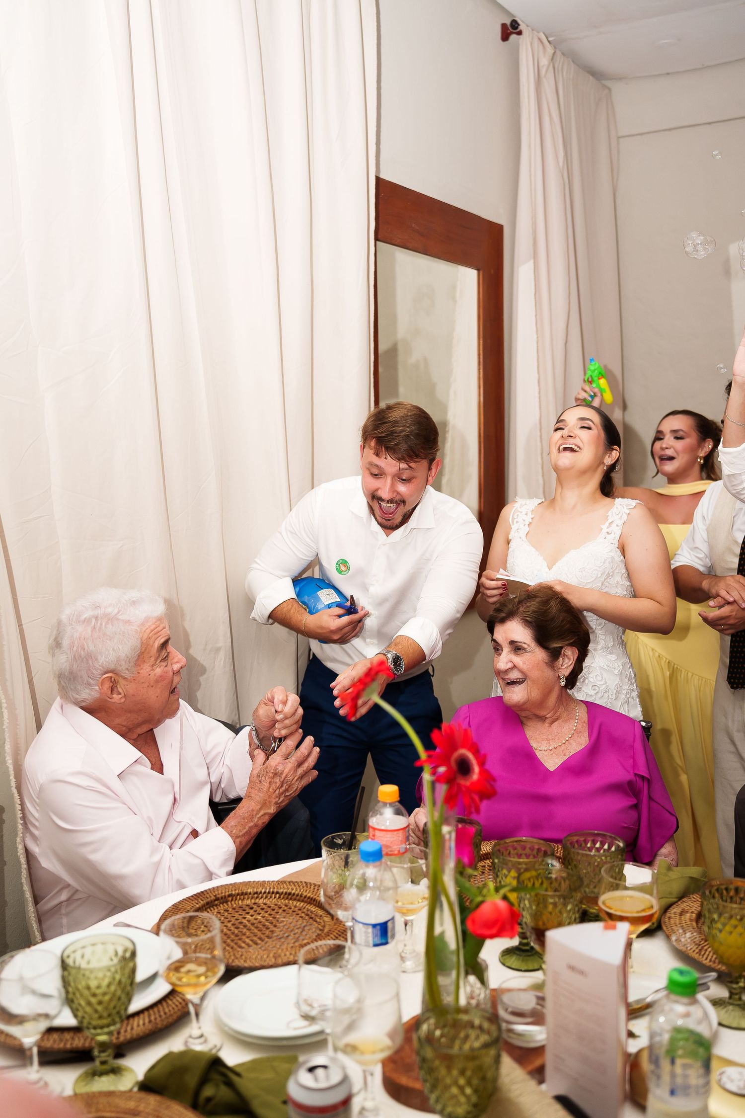 Joyful wedding reception with bride and groom blowing bubbles surrounded by smiling family members at a decorated dinner table.