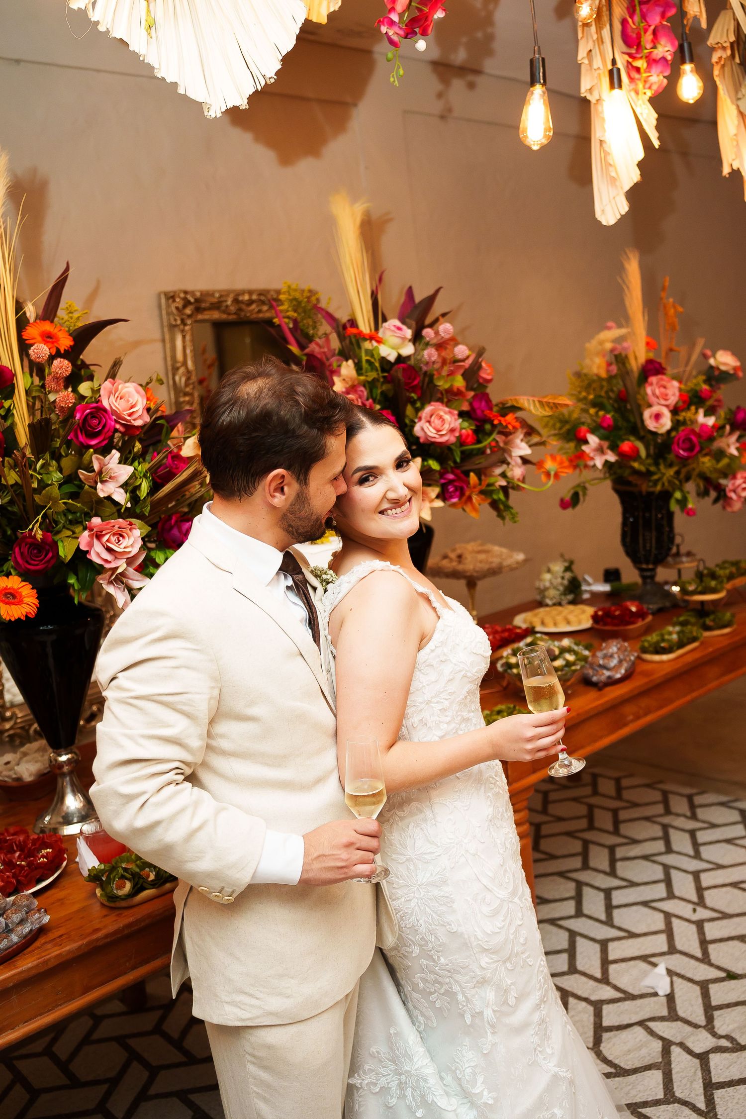 Bride and groom embrace at wedding reception, surrounded by colorful floral arrangements, as bride holds champagne glass.