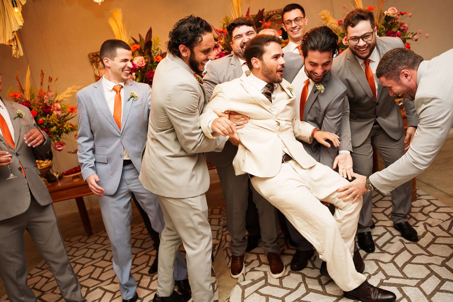 Groomsmen in gray suits with orange ties playfully lift the groom in a cream suit during a lively wedding celebration.
