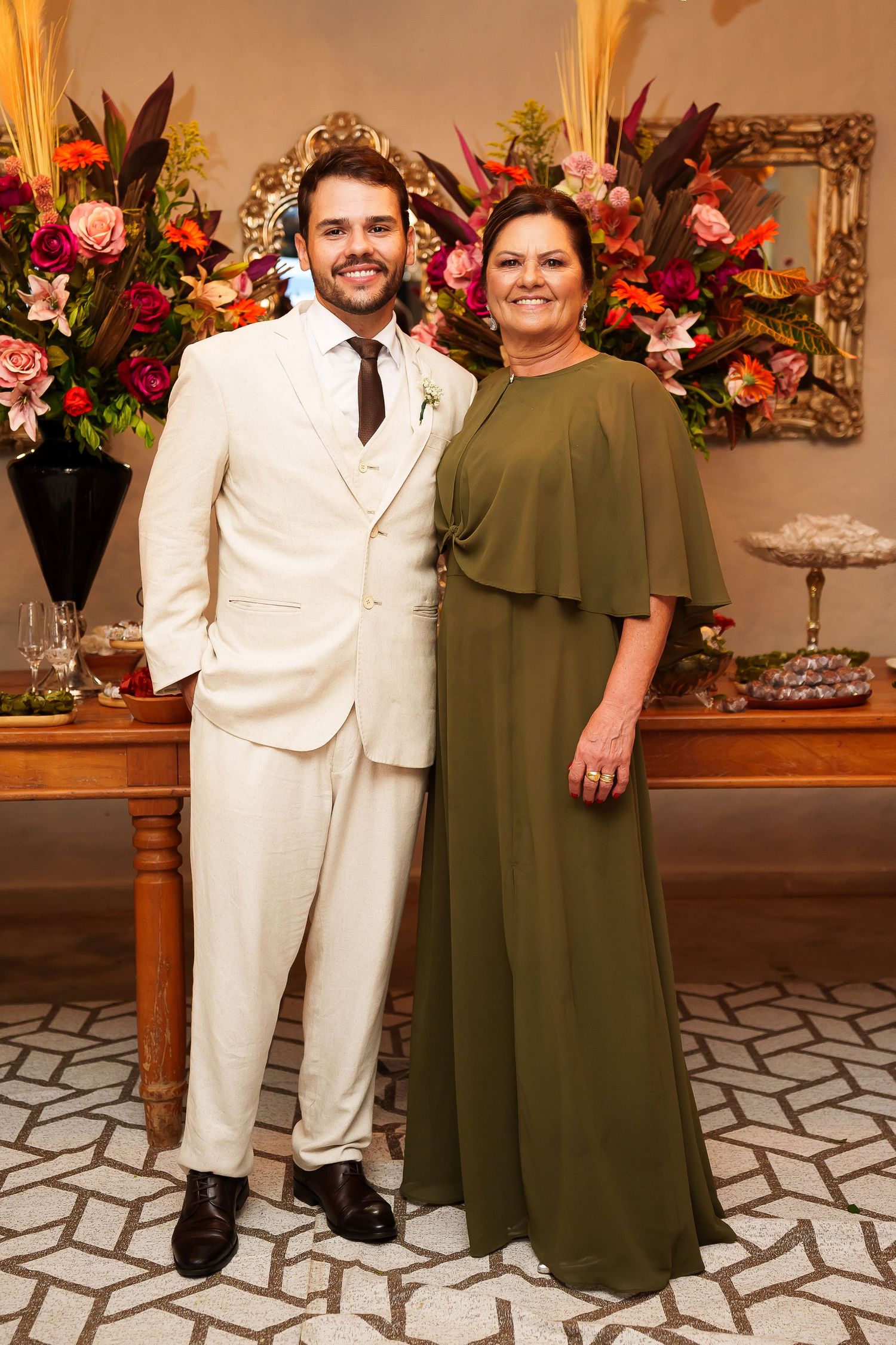 Man in cream suit and woman in olive green gown pose together at elegant floral wedding reception.