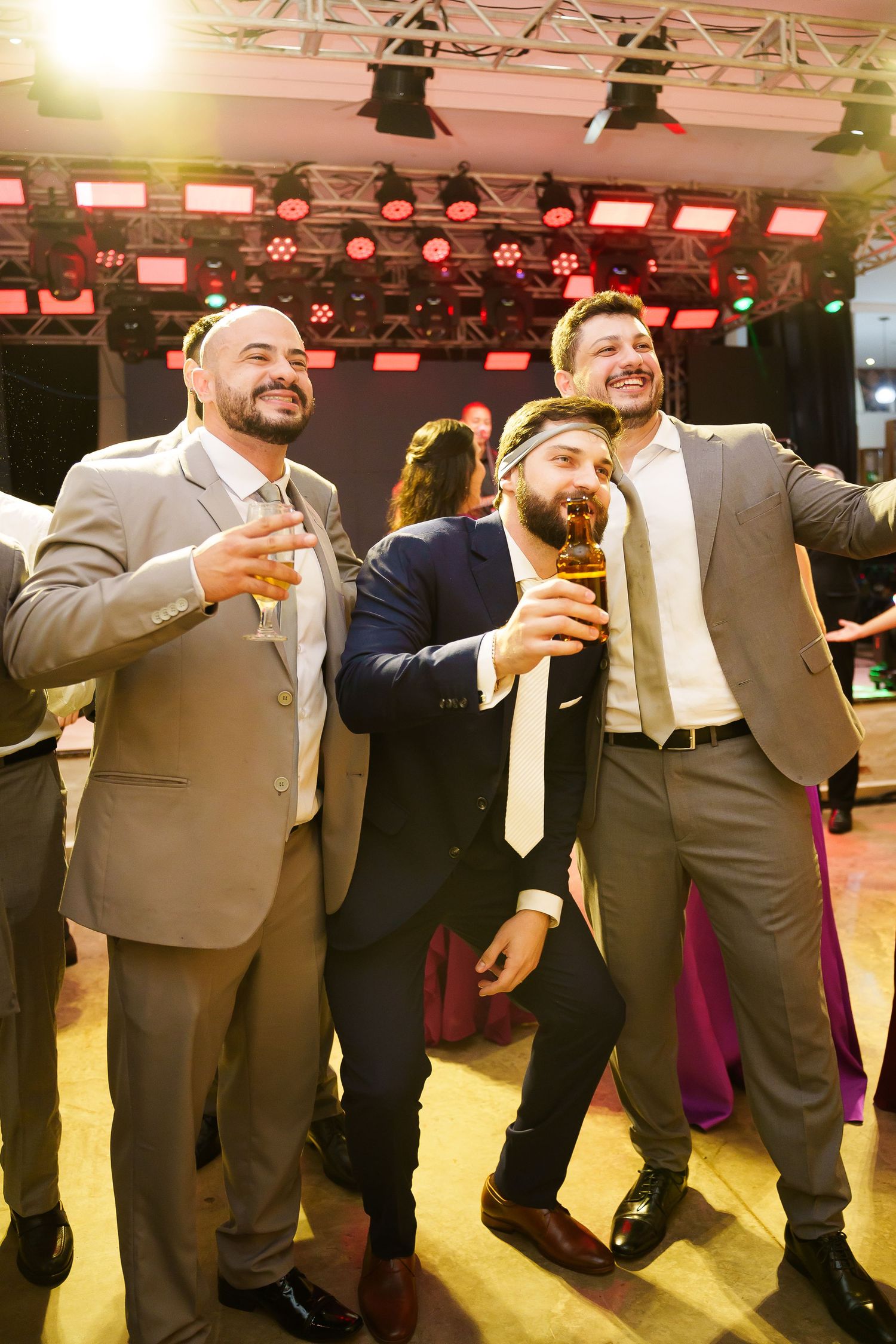 Three men in suits celebrating at a party, holding beer glasses and smiling on a dance floor with stage lights.