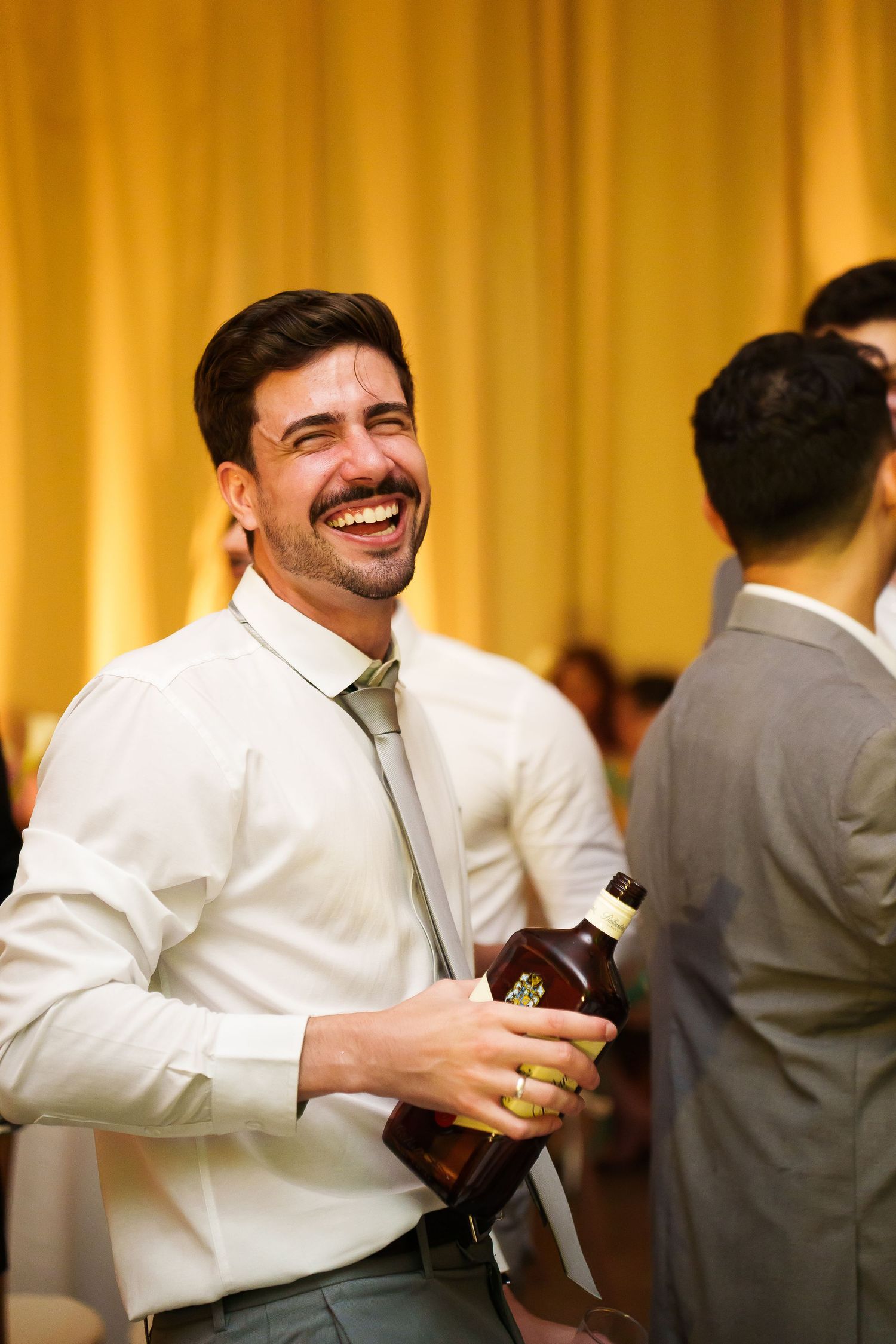 Smiling man in white shirt and gray tie holding a whiskey bottle at a formal event or wedding reception.