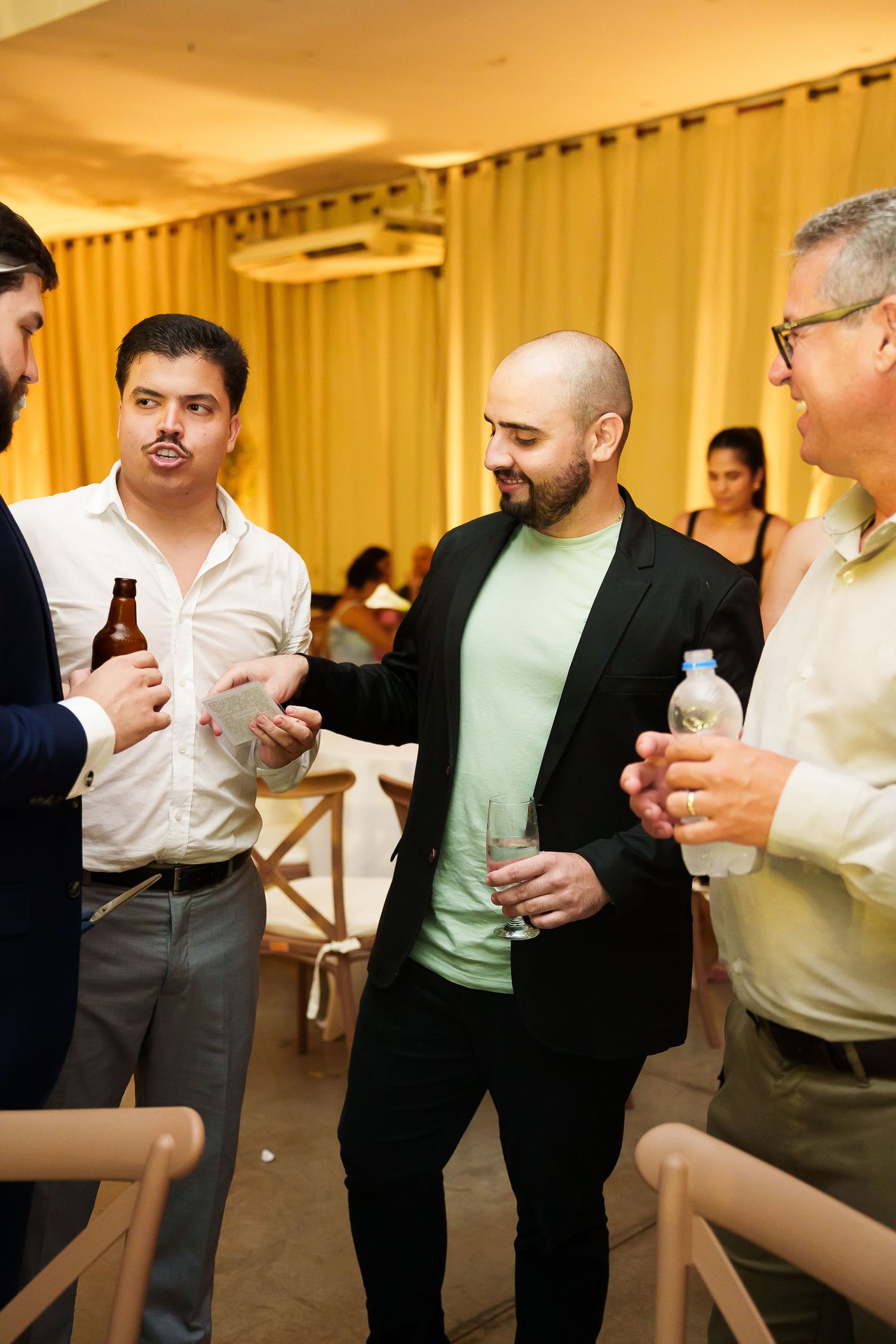 Group of men socializing at an indoor event, holding drinks, wearing smart casual attire in a warmly lit venue.