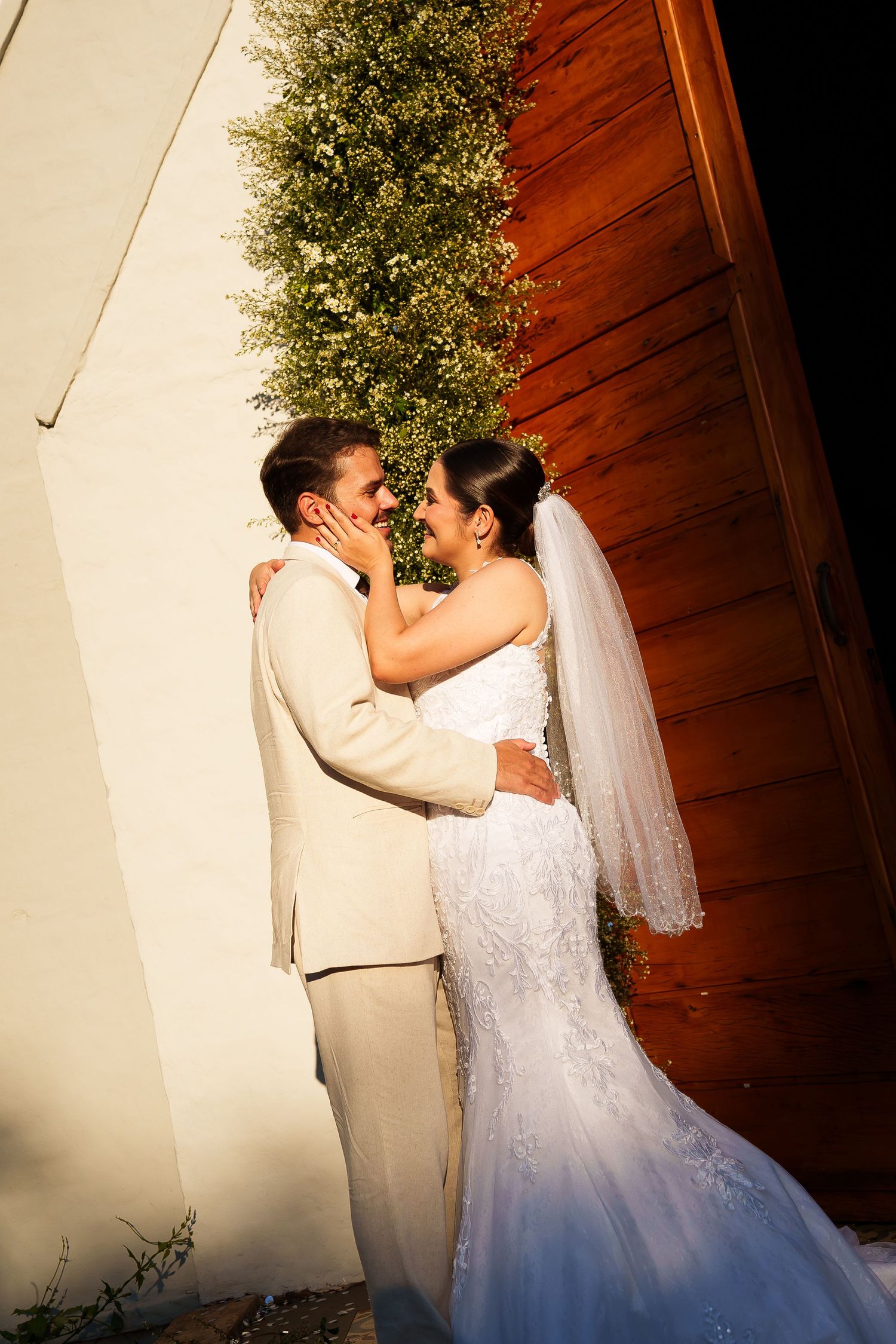 Bride and groom share a romantic moment, embracing and smiling at each other near a white wall and wooden panel outdoors.