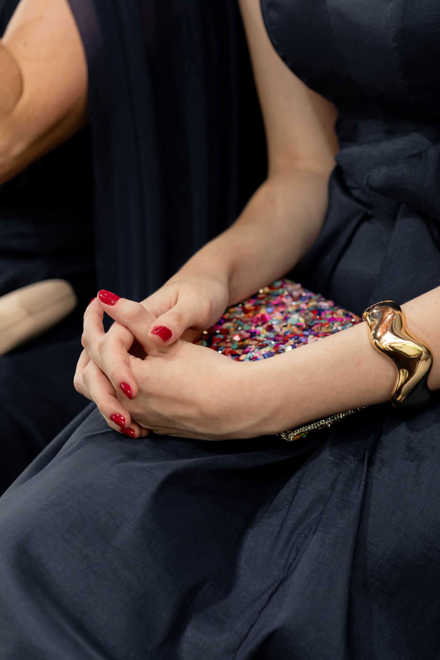 Woman in navy dress with red nails and gold bracelet holds a colorful sequined clutch purse.