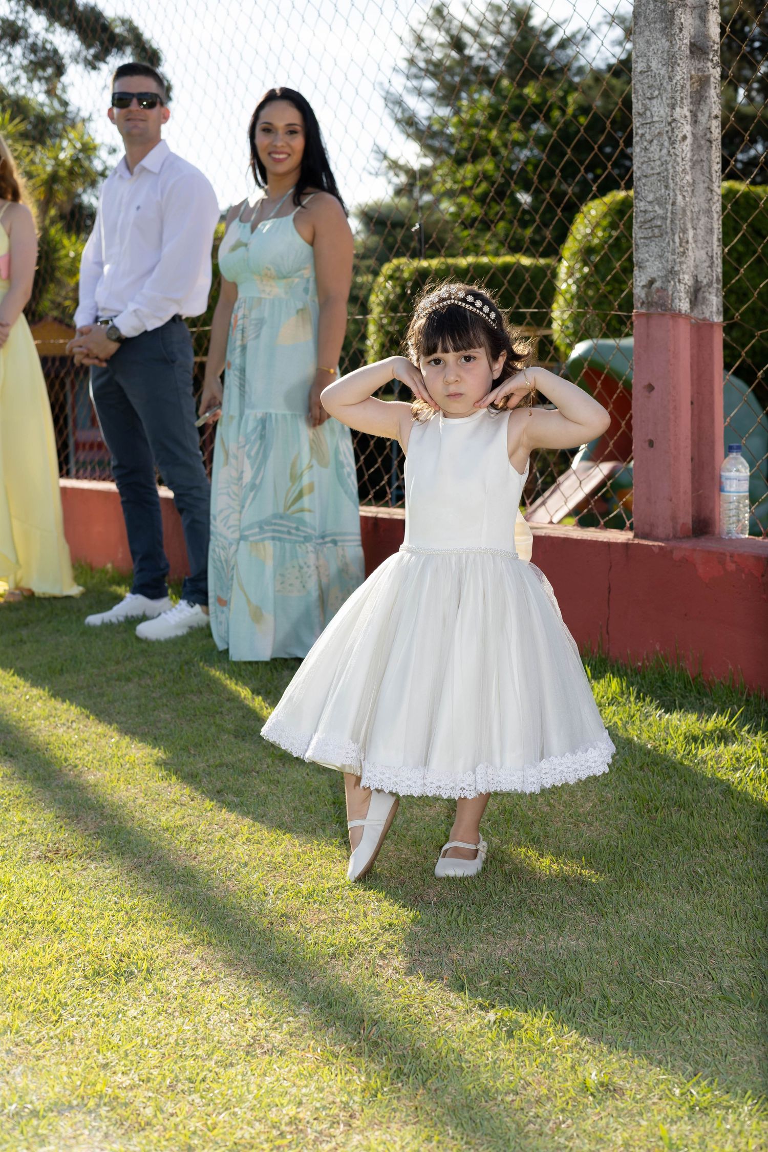 A young girl in a white dress and tiara poses on grass at an outdoor event with adults in formal attire behind her.