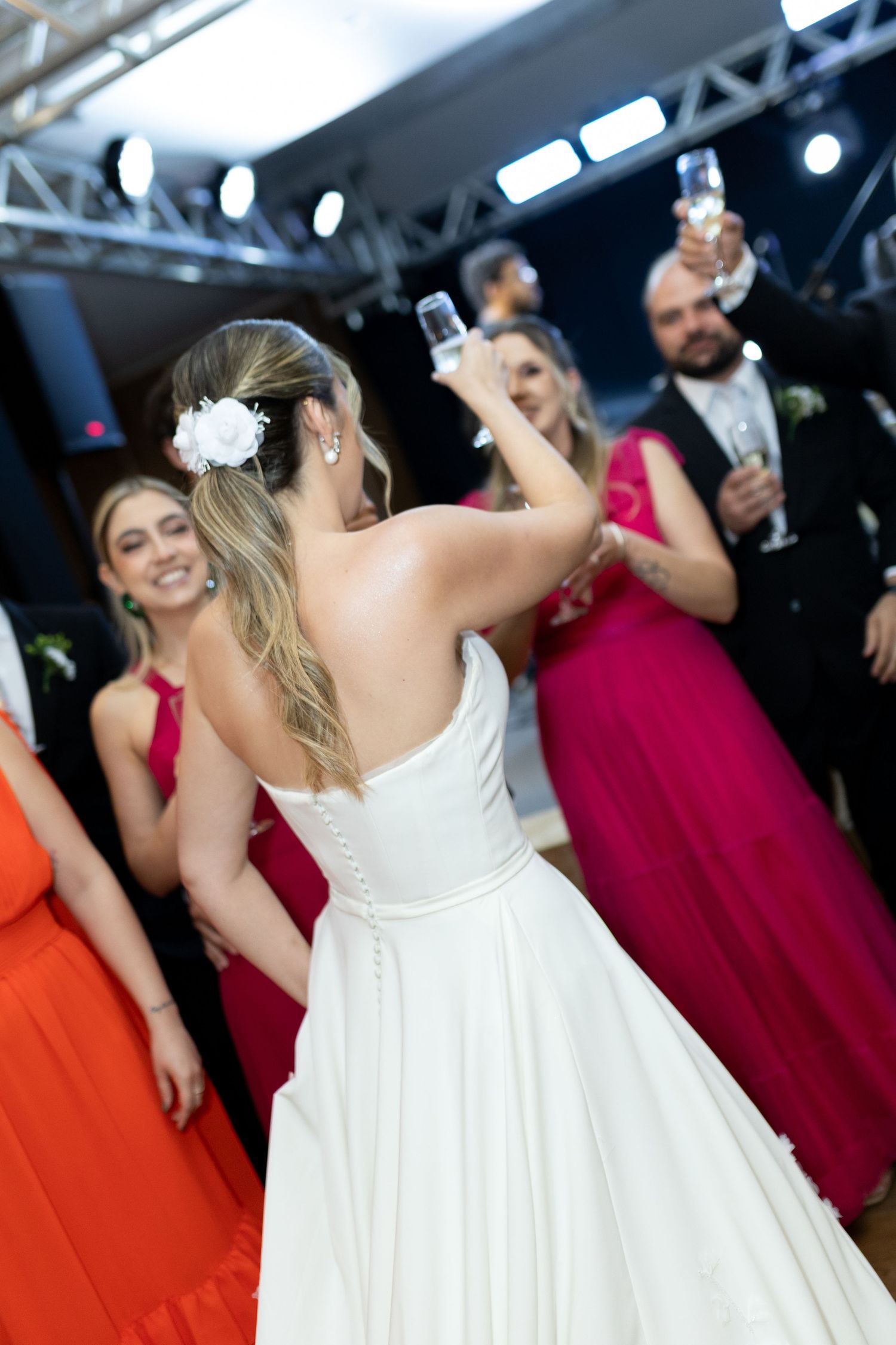 Bride in white gown raises champagne glass with bridesmaids in red and pink dresses during wedding reception toast.