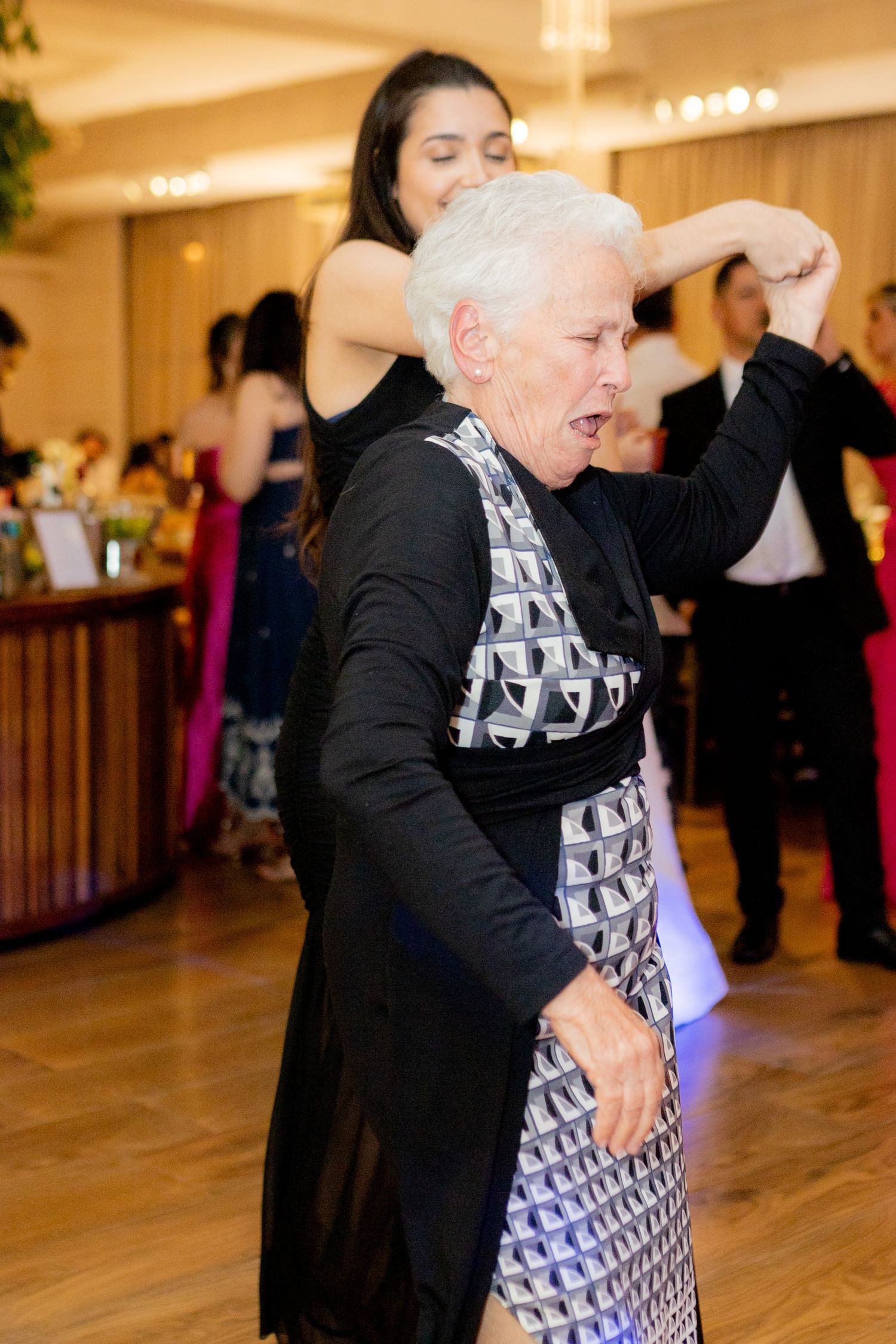Elderly woman in black and silver dress dancing joyfully at a formal event with guests in background.