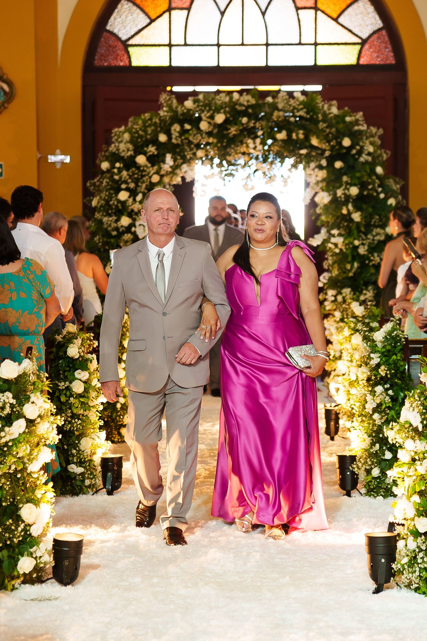 Couple walks down floral wedding aisle, man in tan suit, woman in magenta gown, white flower arch backdrop.