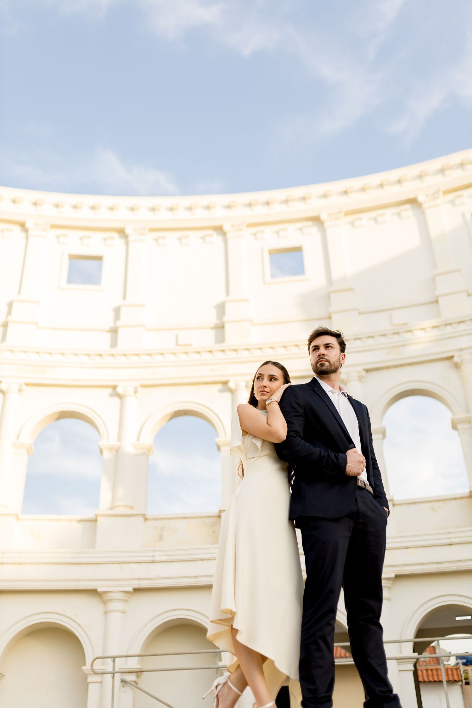 Elegant couple posing in front of a grand Roman colosseum-style architecture under a blue sky.