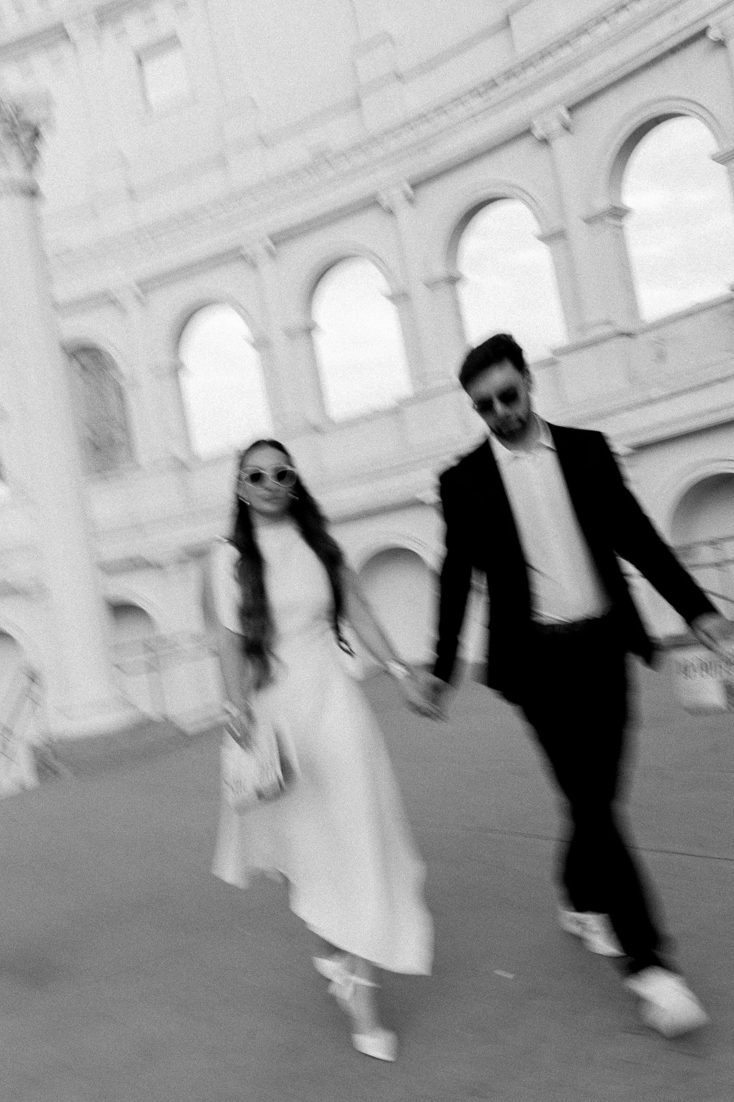 Black and white photo of a couple holding hands walking near the Colosseum in Rome, wearing sunglasses and formal attire.