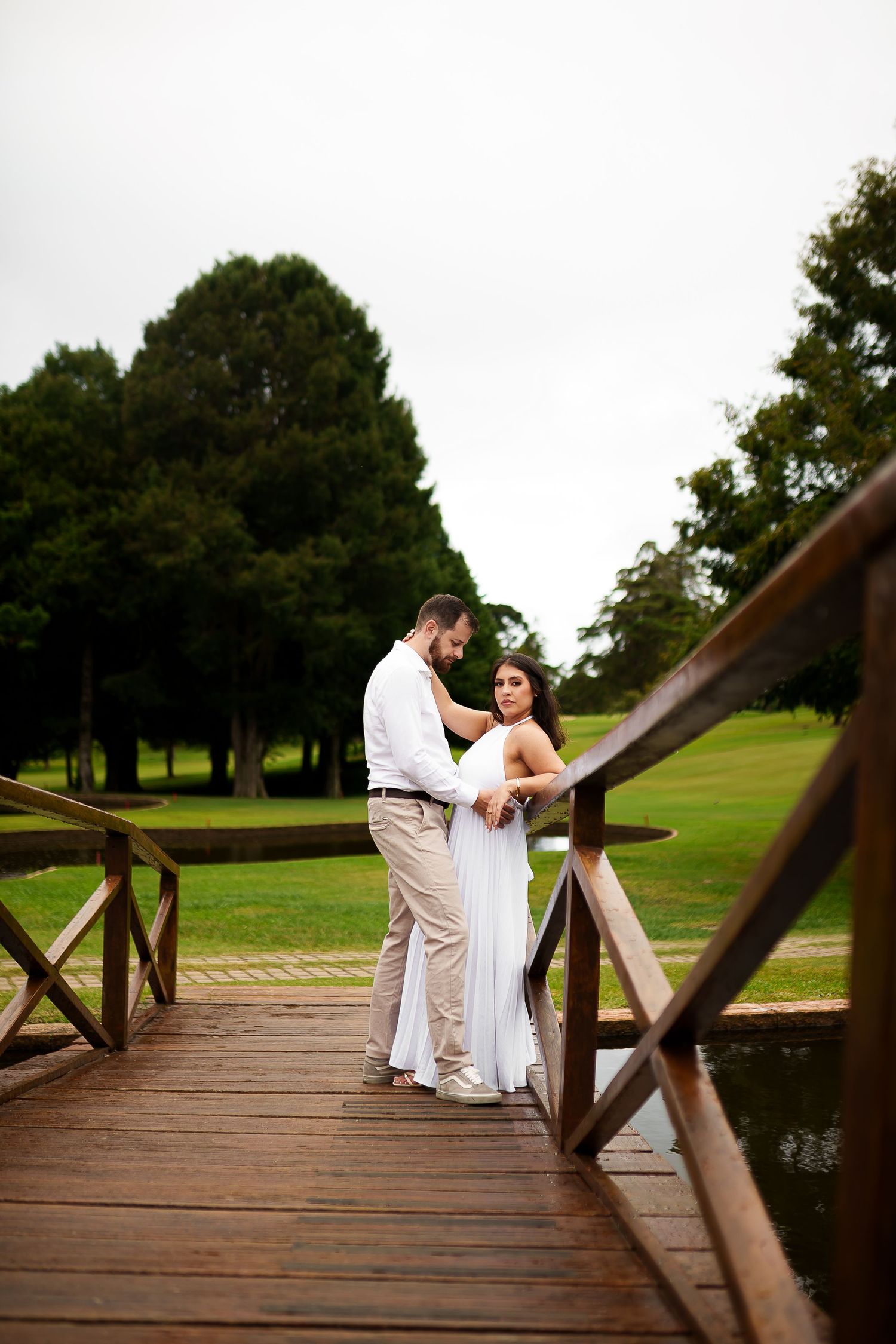 Couple in wedding attire embracing on a wooden bridge surrounded by lush green trees and grass on a cloudy day.