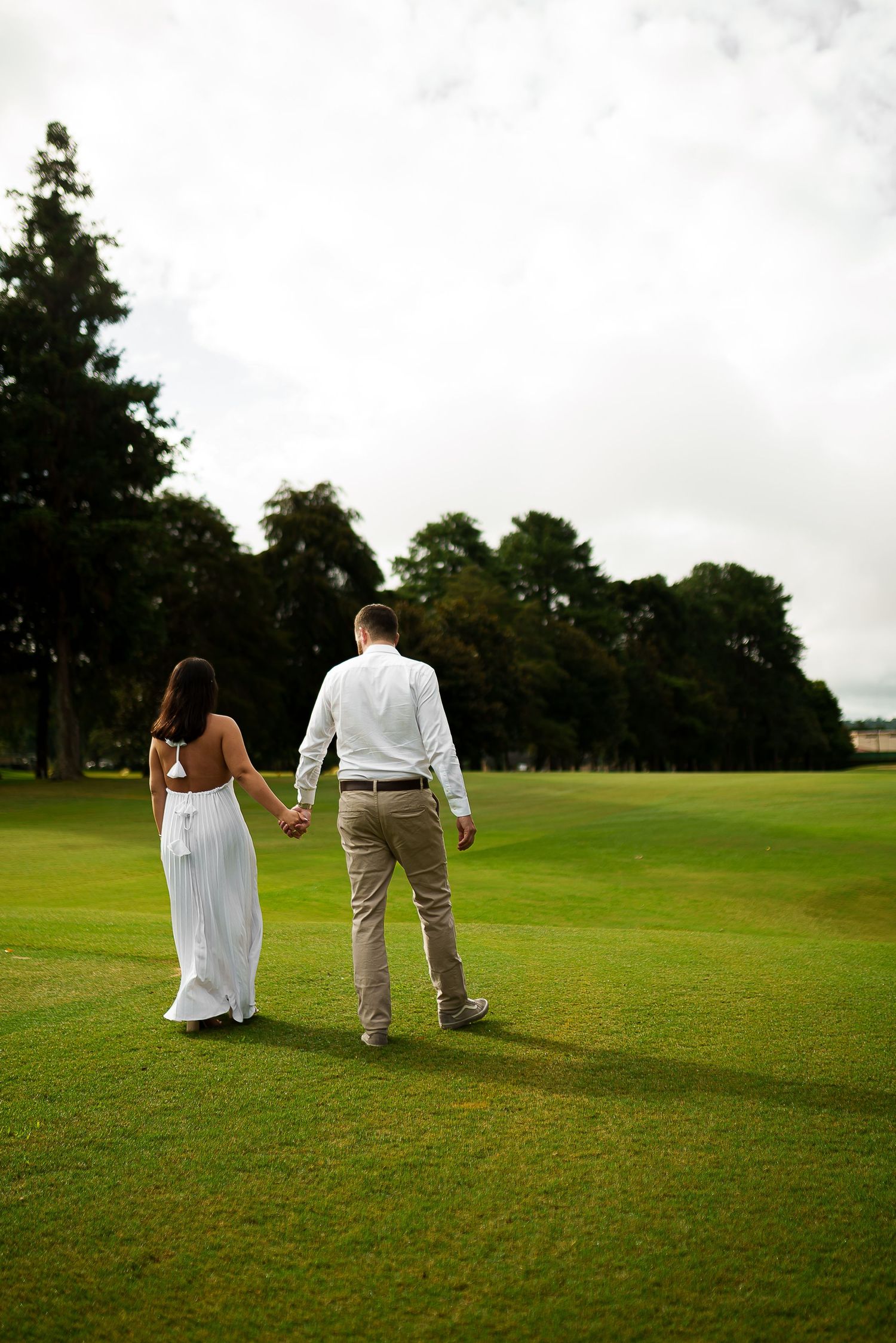 Couple in white attire holding hands while walking on lush green grass with trees in background.