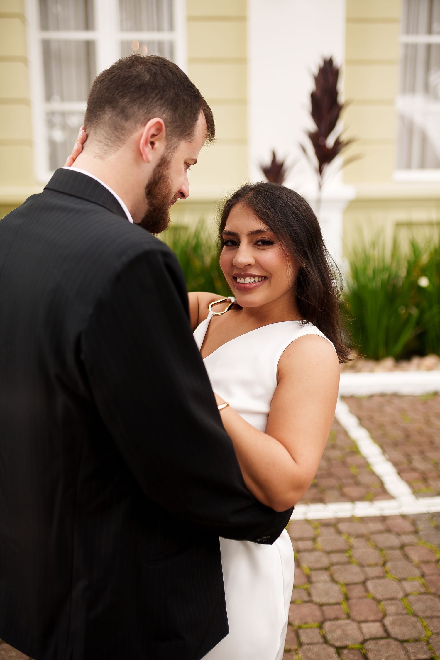 A smiling bride in a white dress embraces her groom in a black suit outdoors near a yellow building.