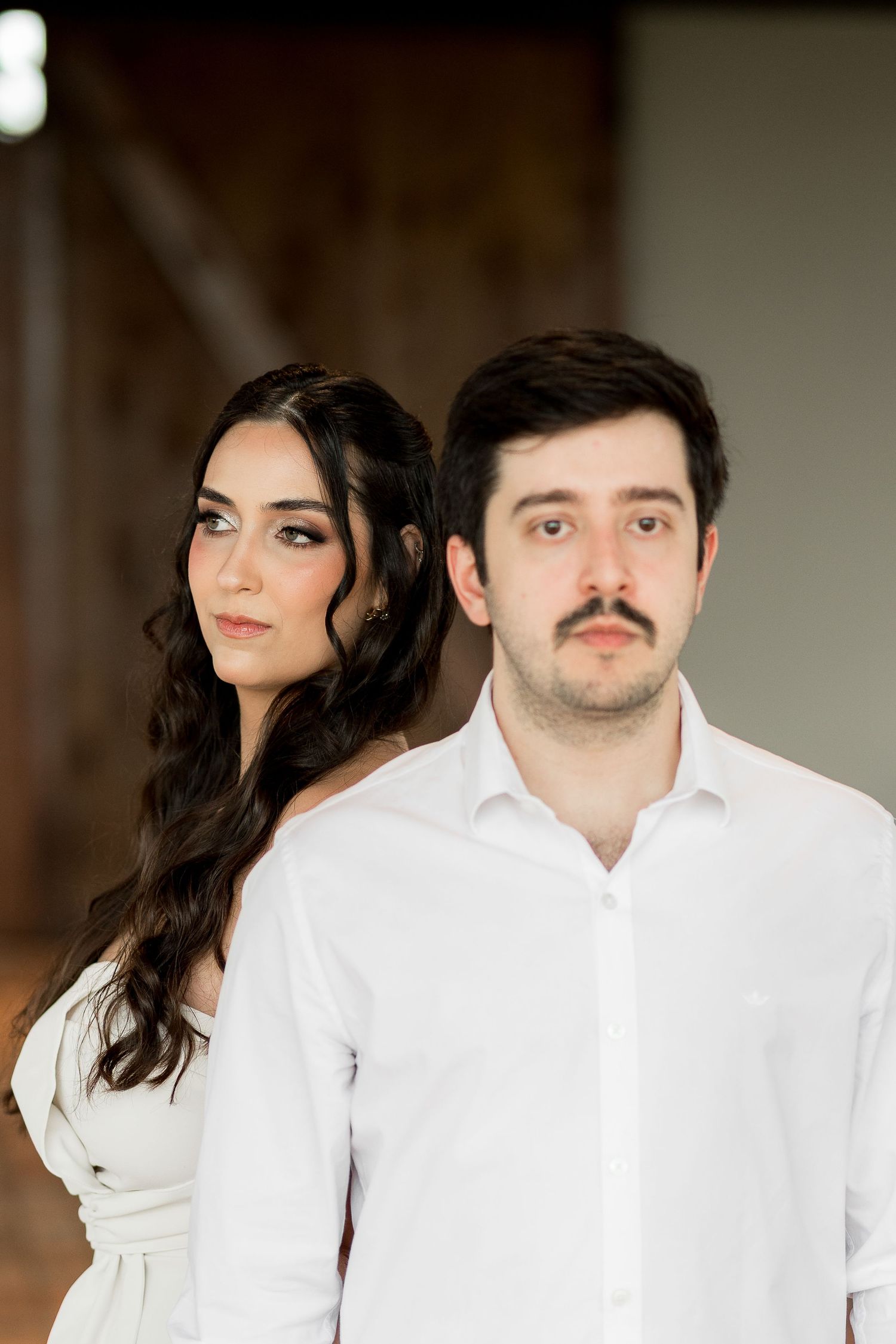 A young couple in white attire poses together, the woman with long dark hair and makeup, the man with a mustache.