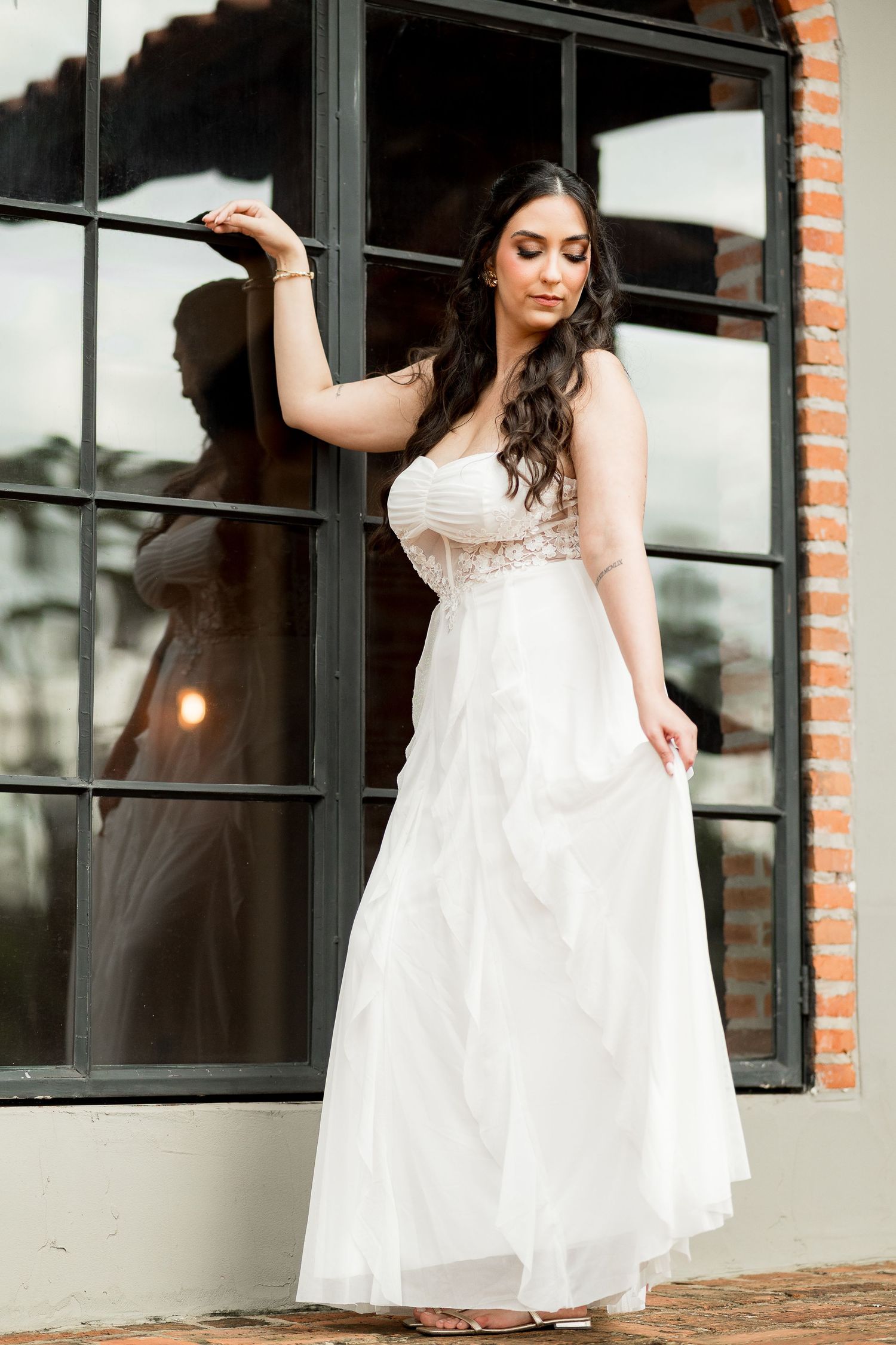 Bride in white strapless lace gown poses elegantly by black-framed windows with brick wall backdrop.