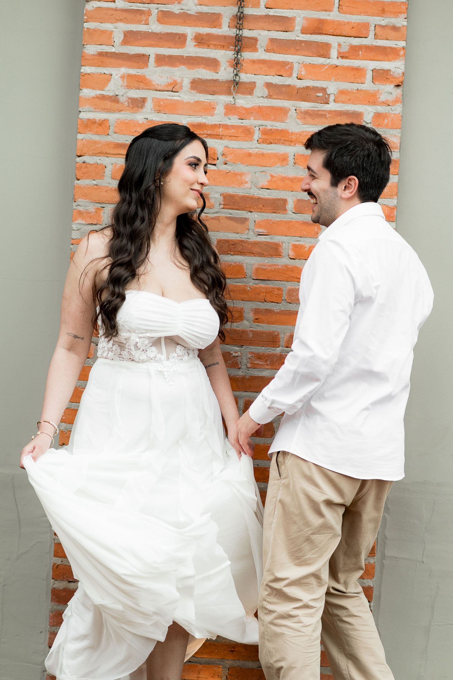 A couple in white wedding attire smiling at each other while holding hands in front of a rustic brick wall.