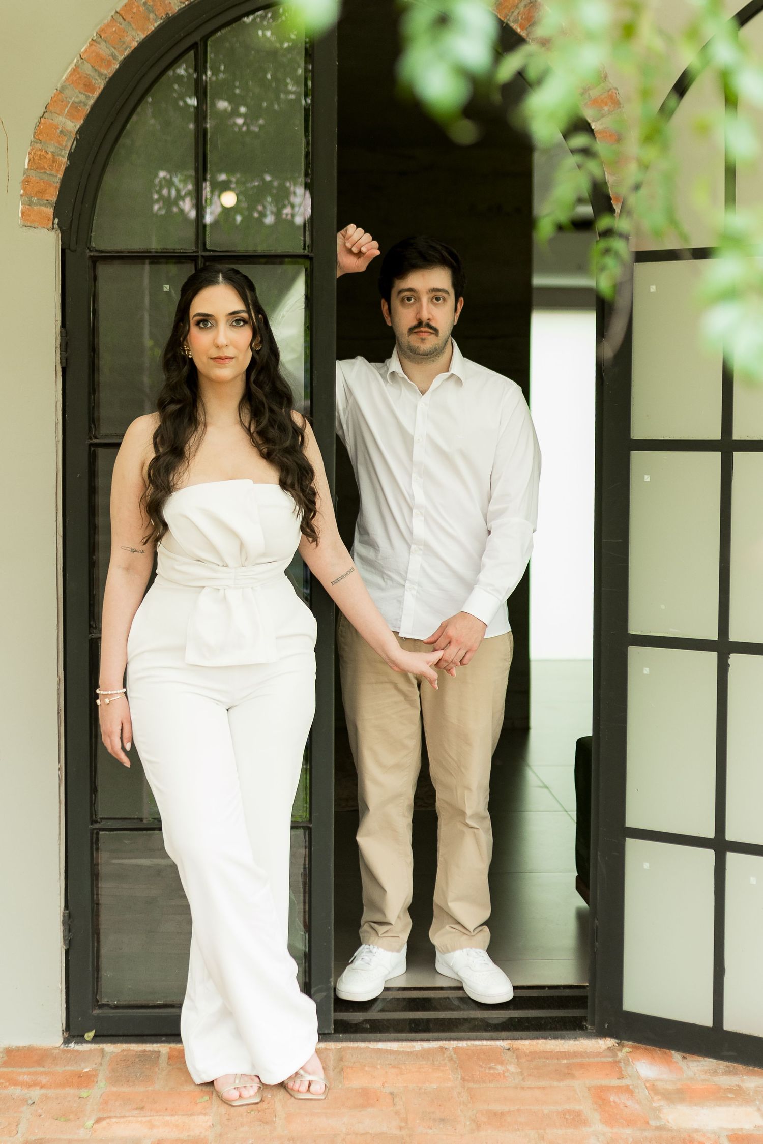 Couple in white outfits posing at an arched black French door, woman in jumpsuit and man in shirt holding hands.