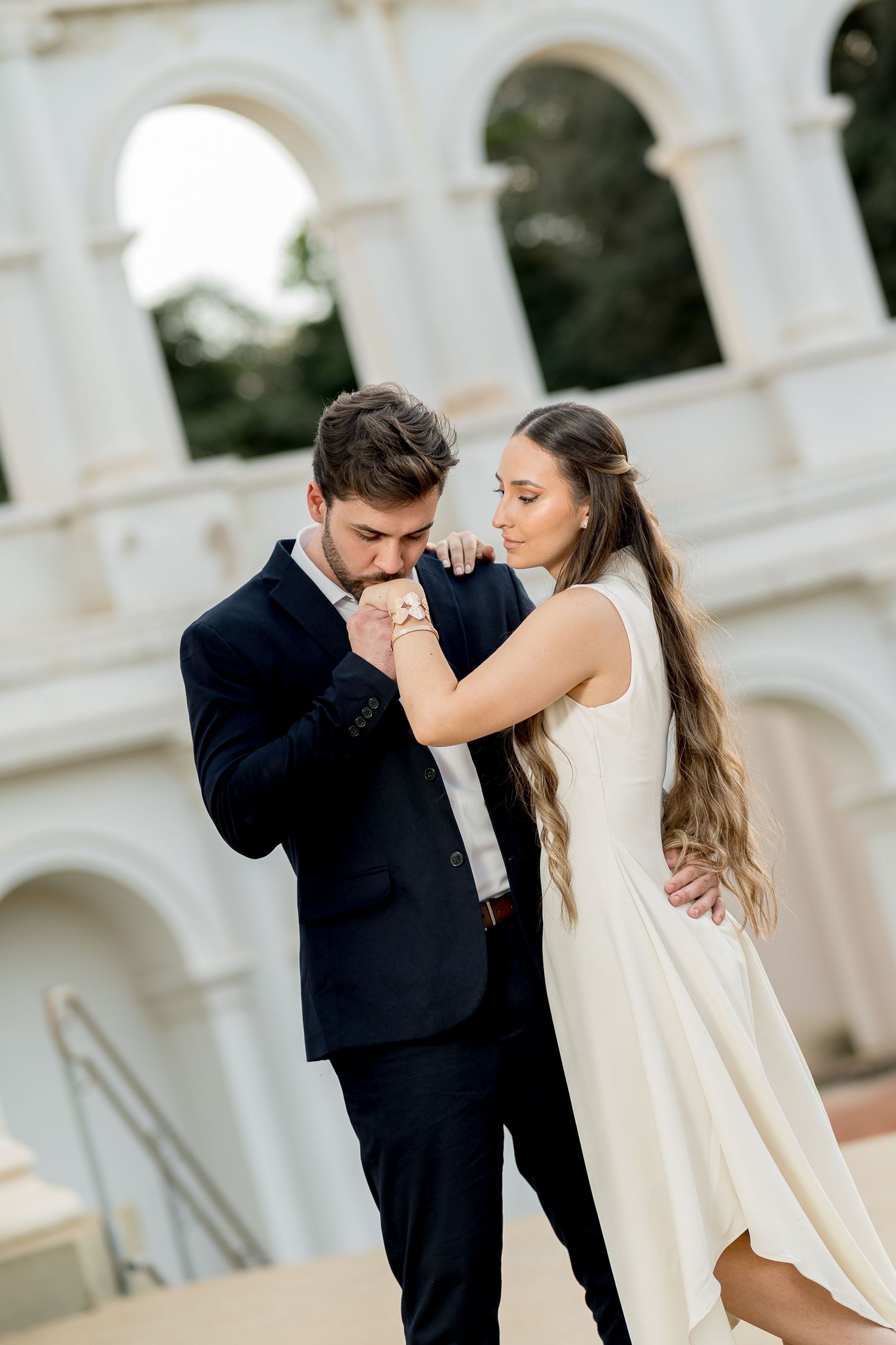Groom in navy suit kissing bride's hand outdoors near elegant arched building during romantic wedding photoshoot.