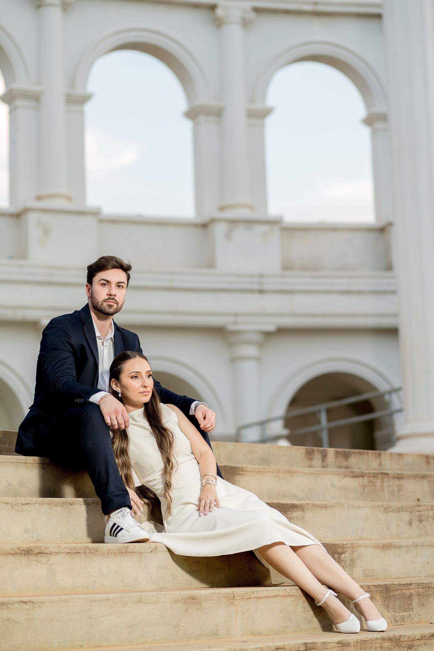 Elegant couple sitting on stone steps before classical arched building, woman in white dress, man in dark suit.