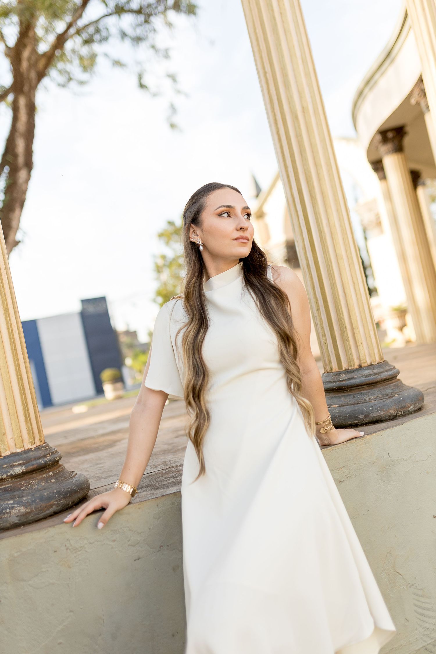 Young woman in white dress poses gracefully between classical columns outdoors, her long hair flowing elegantly.