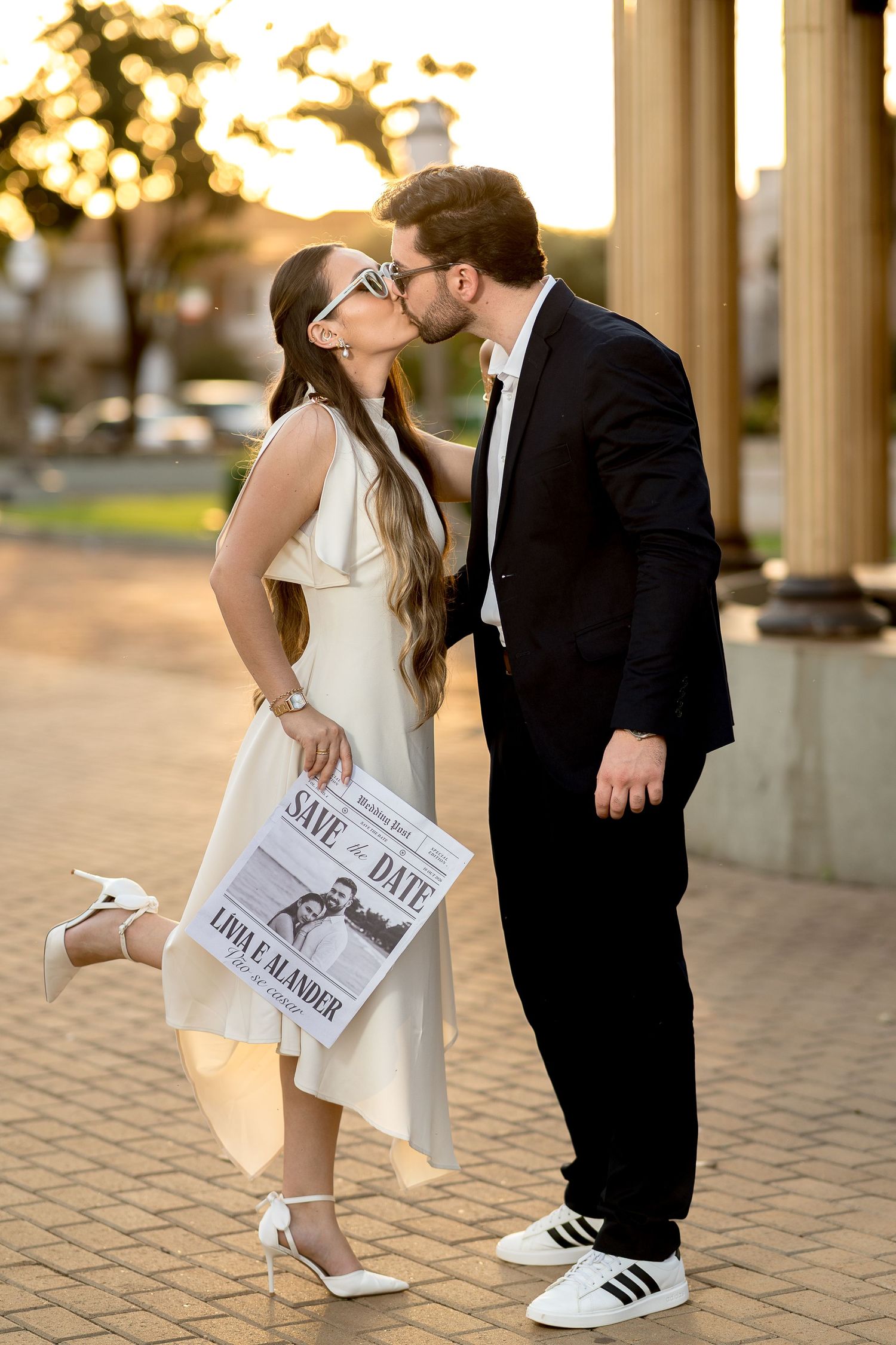 Couple kissing outdoors at golden hour, woman in white dress holding Save the Date newspaper, man in black suit.