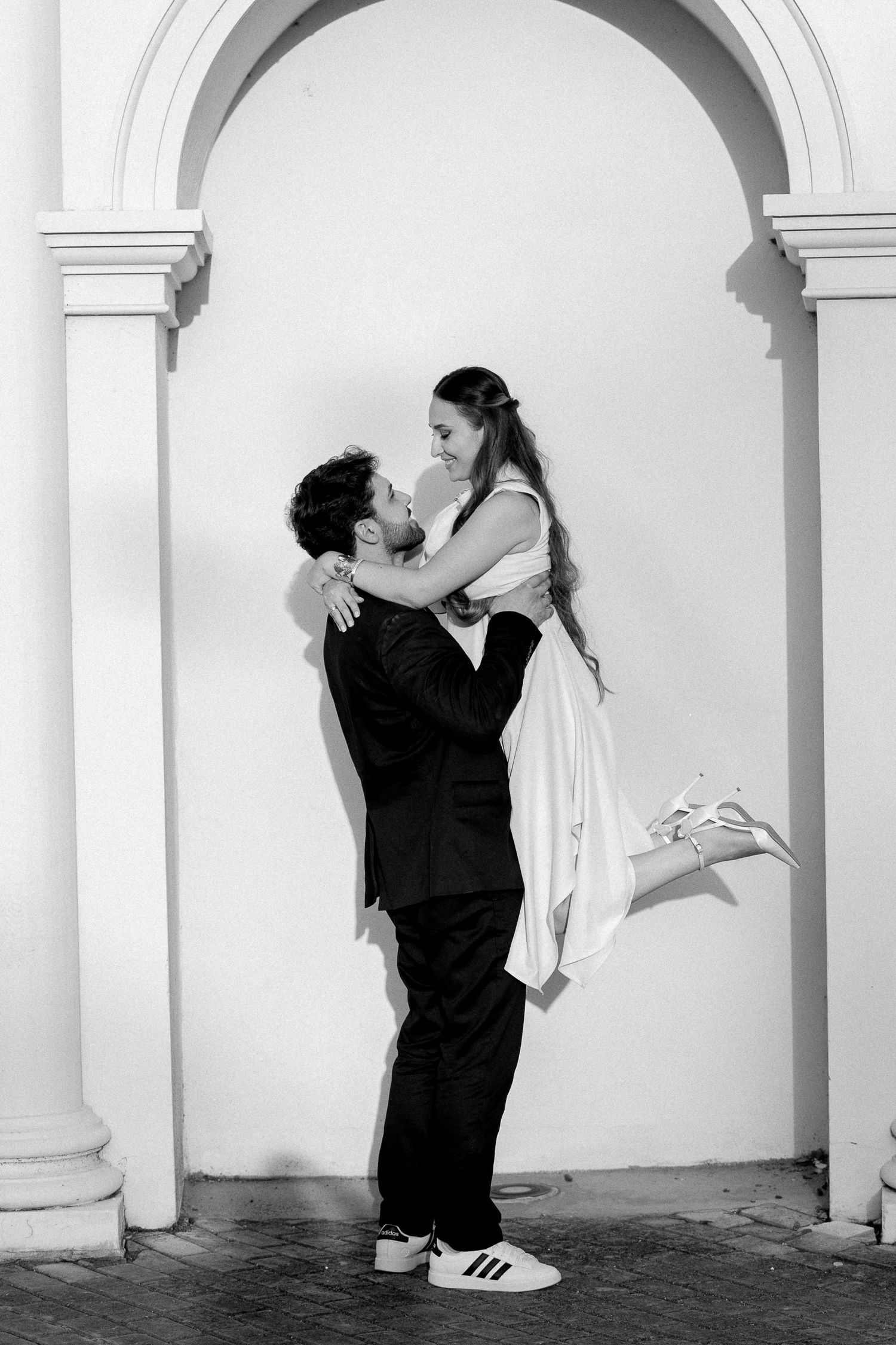Black and white photo of groom lifting bride in wedding dress near elegant arch, romantic outdoor portrait.