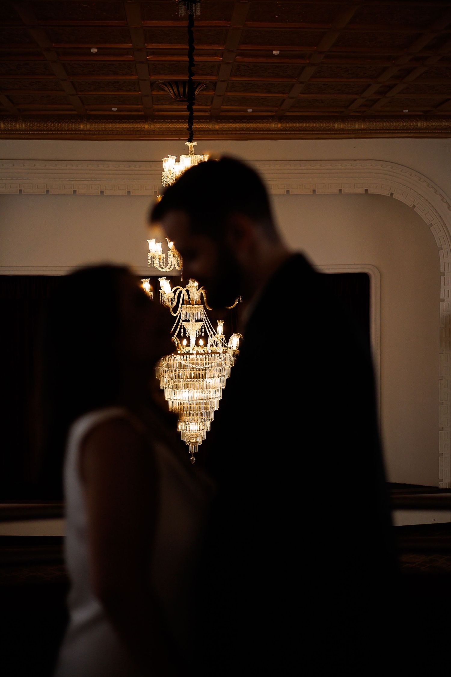 Silhouetted couple sharing romantic moment in elegant ballroom with glowing crystal chandelier backdrop.