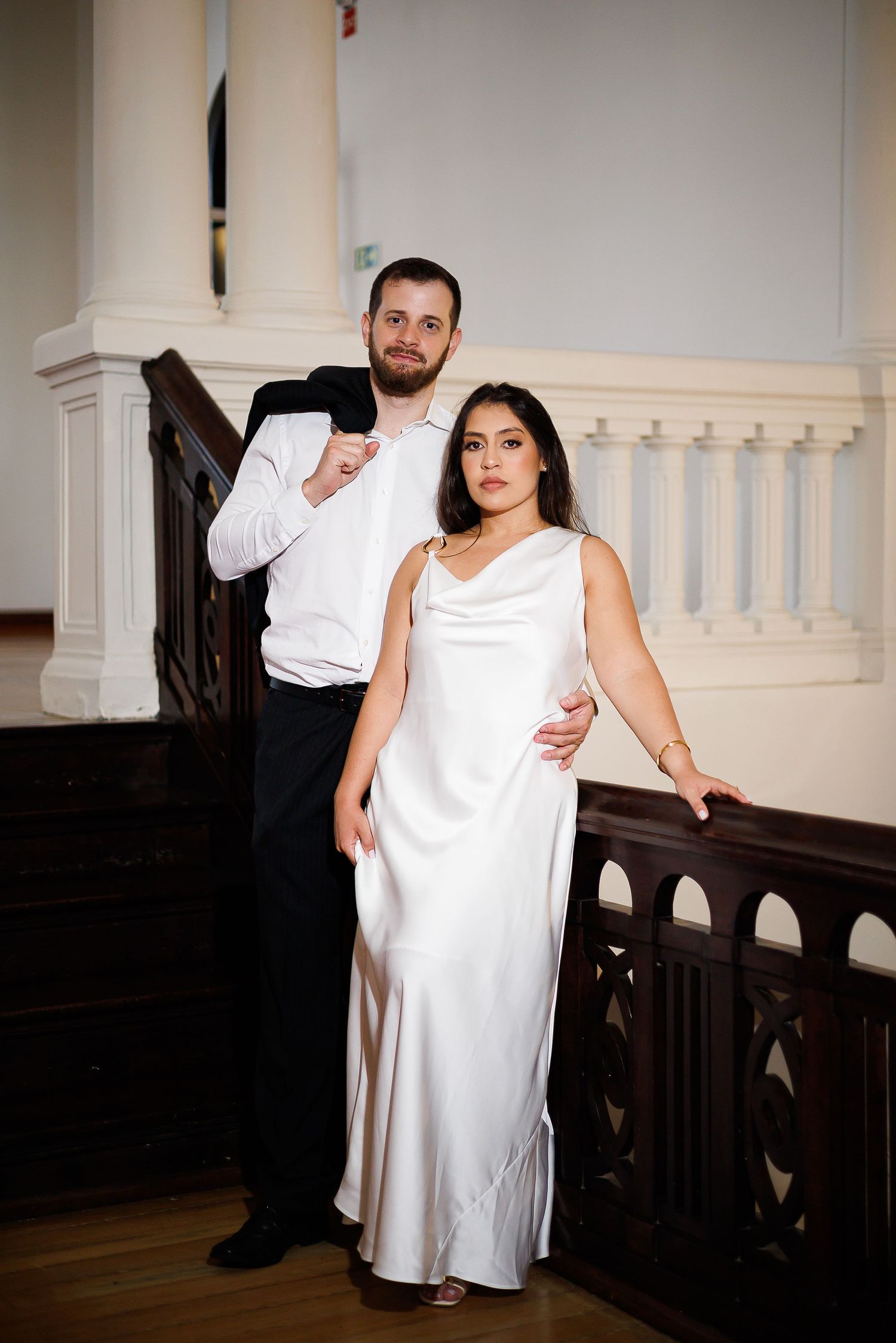 Elegant couple posing on grand staircase, woman in white satin gown, man in black suit, formal venue.