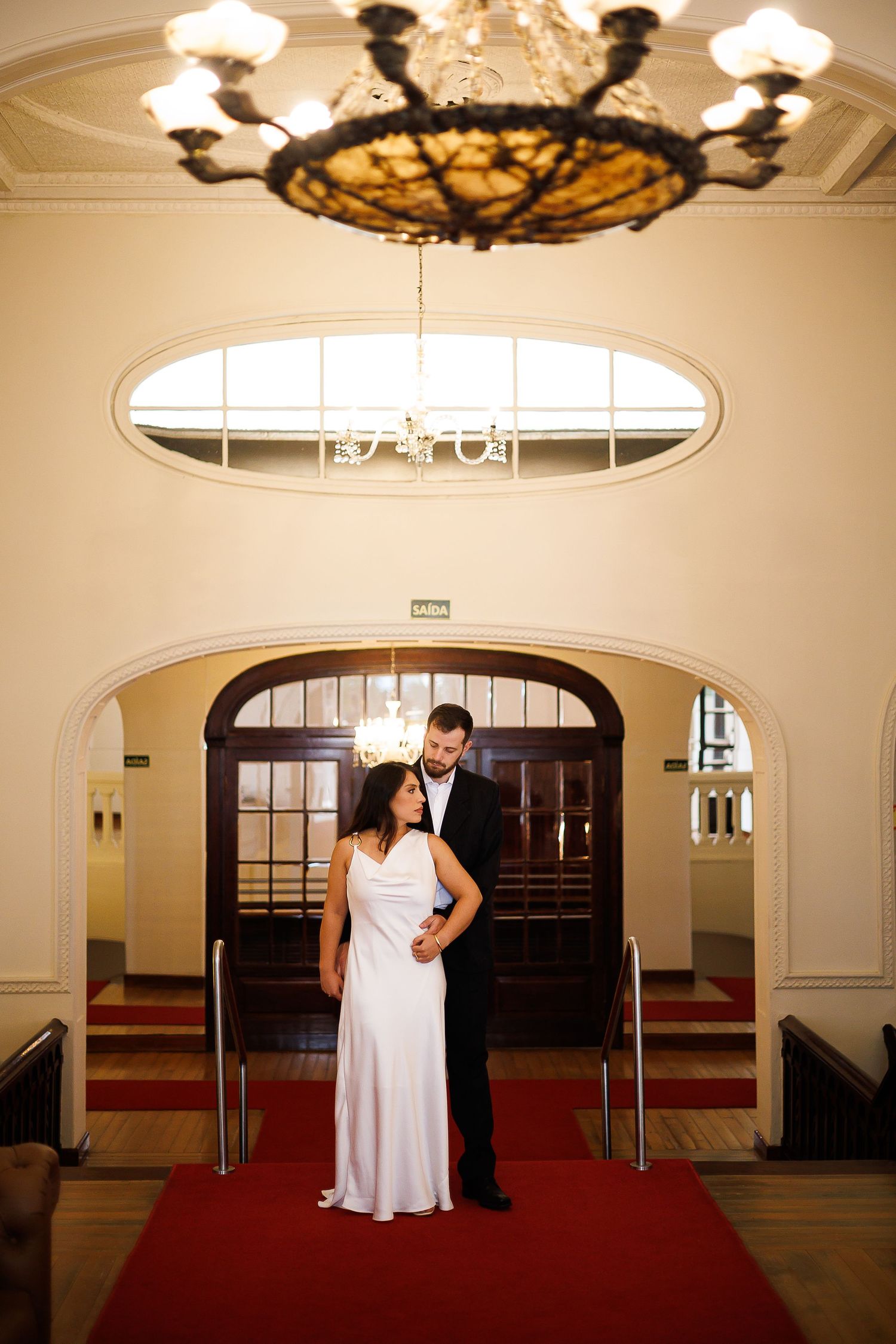 Bride in white gown and groom in tuxedo stand on red carpet under elegant chandelier in grand venue.