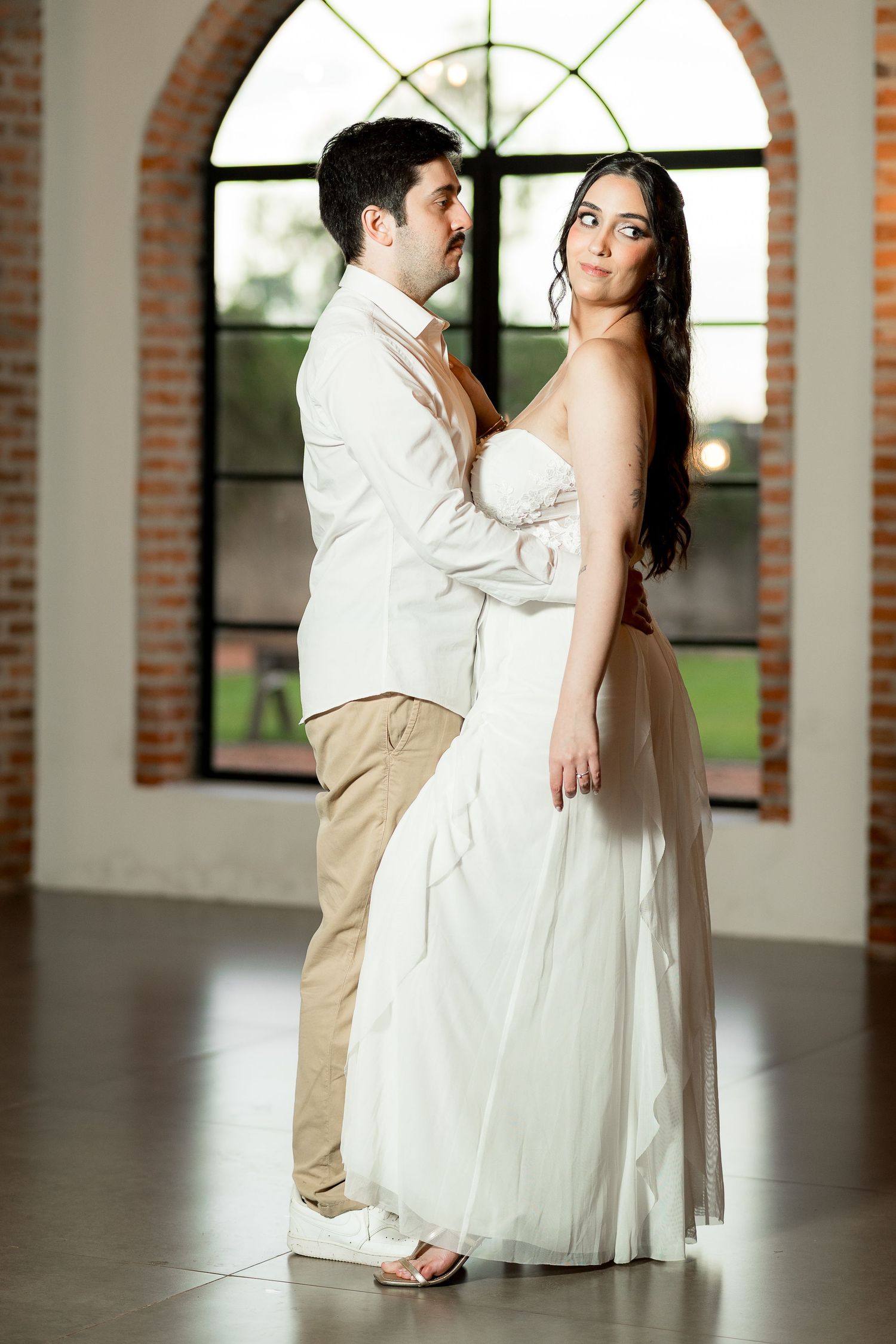 Elegant couple in wedding attire posing in front of arched brick window, bride in white gown, groom in white jacket.